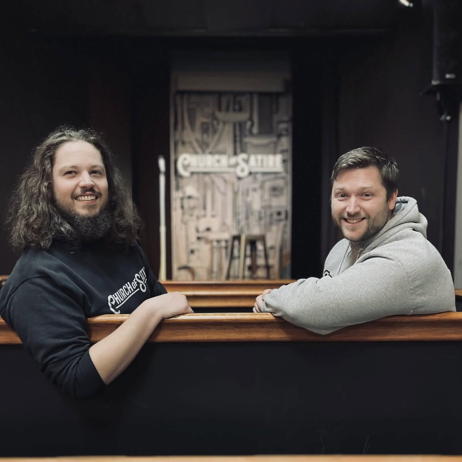 Two men sitting at a bar counter smiling, with a mural in the background, one with long curly hair and the other with short dark hair, both wearing casual clothing.