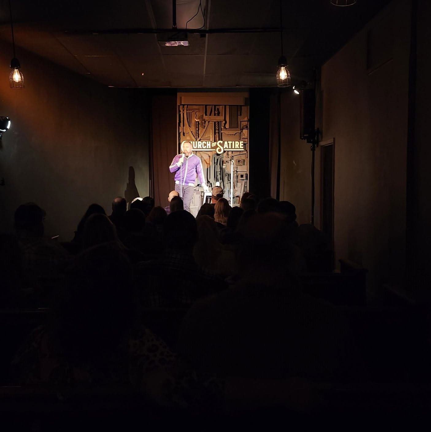 Person on stage performing in a dark room with audience, illuminated by stage lights, with a sign reading 'Church Satire' behind.