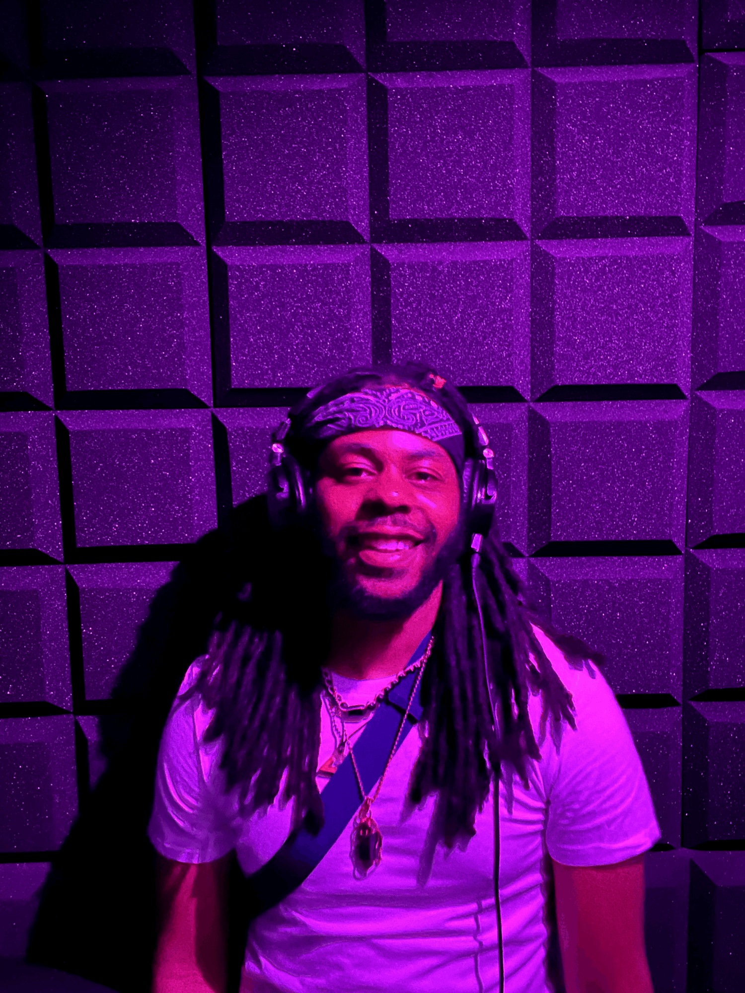 Person with long dreadlocks wearing headphones and a white shirt, smiling, in a purple-lit soundproof room.
