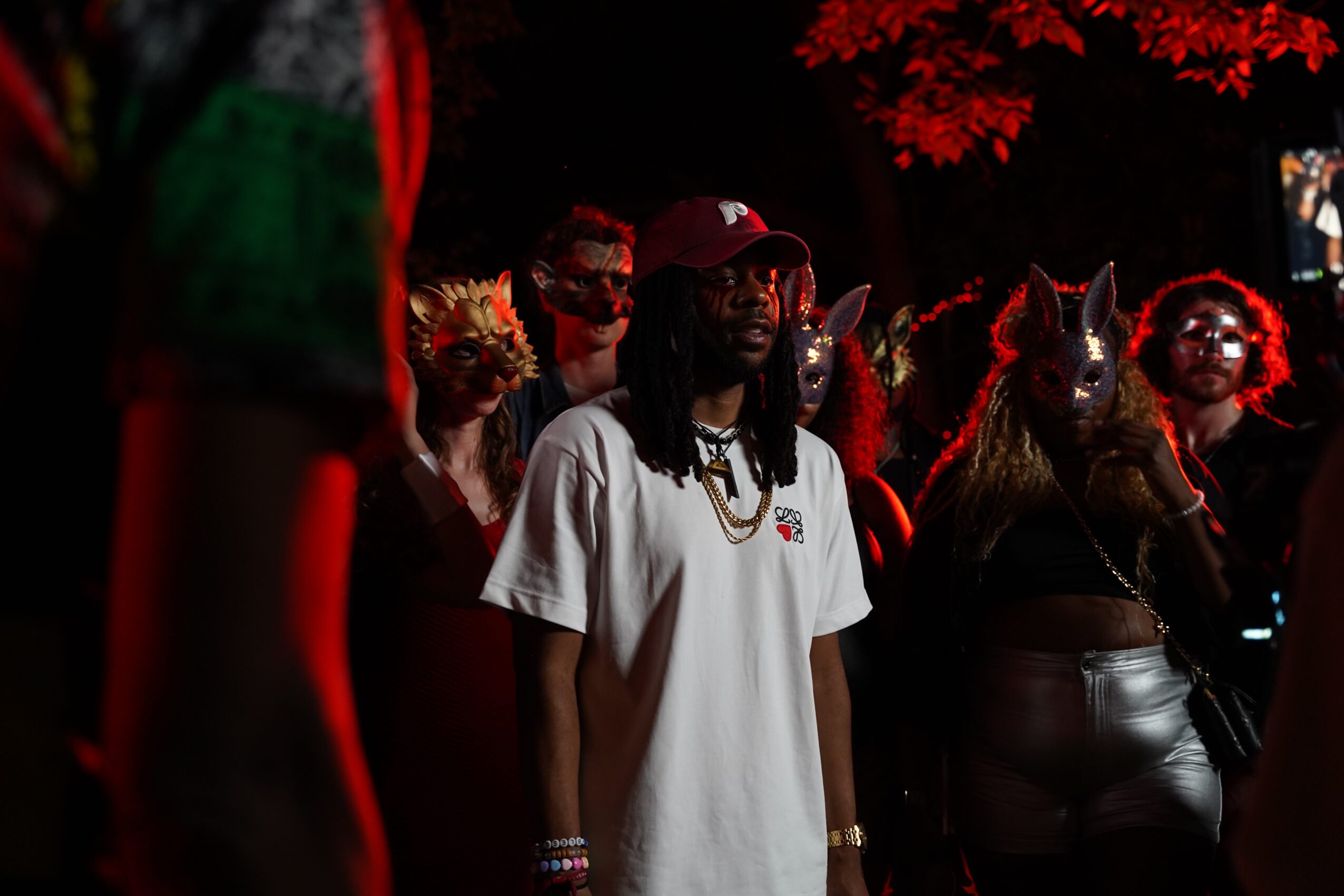 Group of people, some wearing masks and costumes, standing outdoors at night with red leaves in the background.