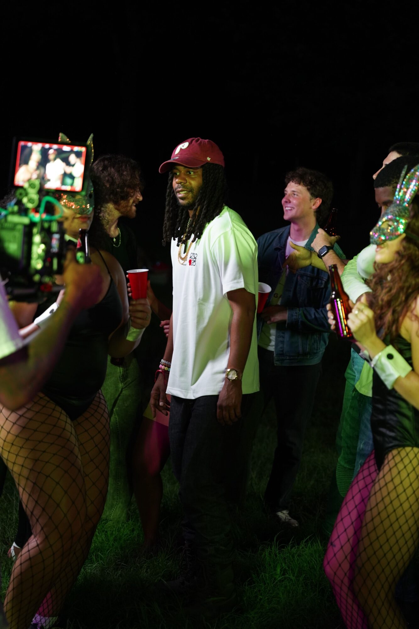 Group of people at night, some holding drinks, with a person in a white shirt in the center, and a person with a phone in the background.