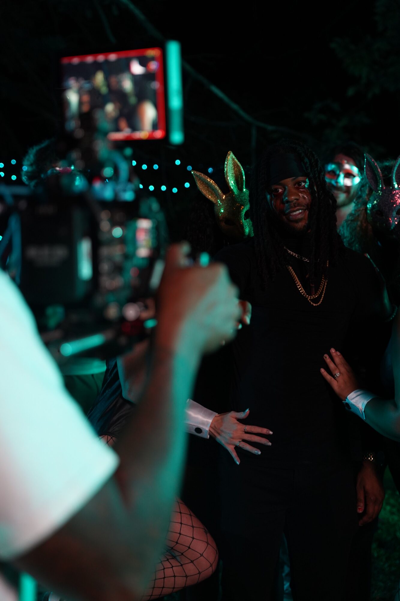Person operating camera in dark environment with people and equipment visible, some with glasses, hands reaching out, colorful lights.