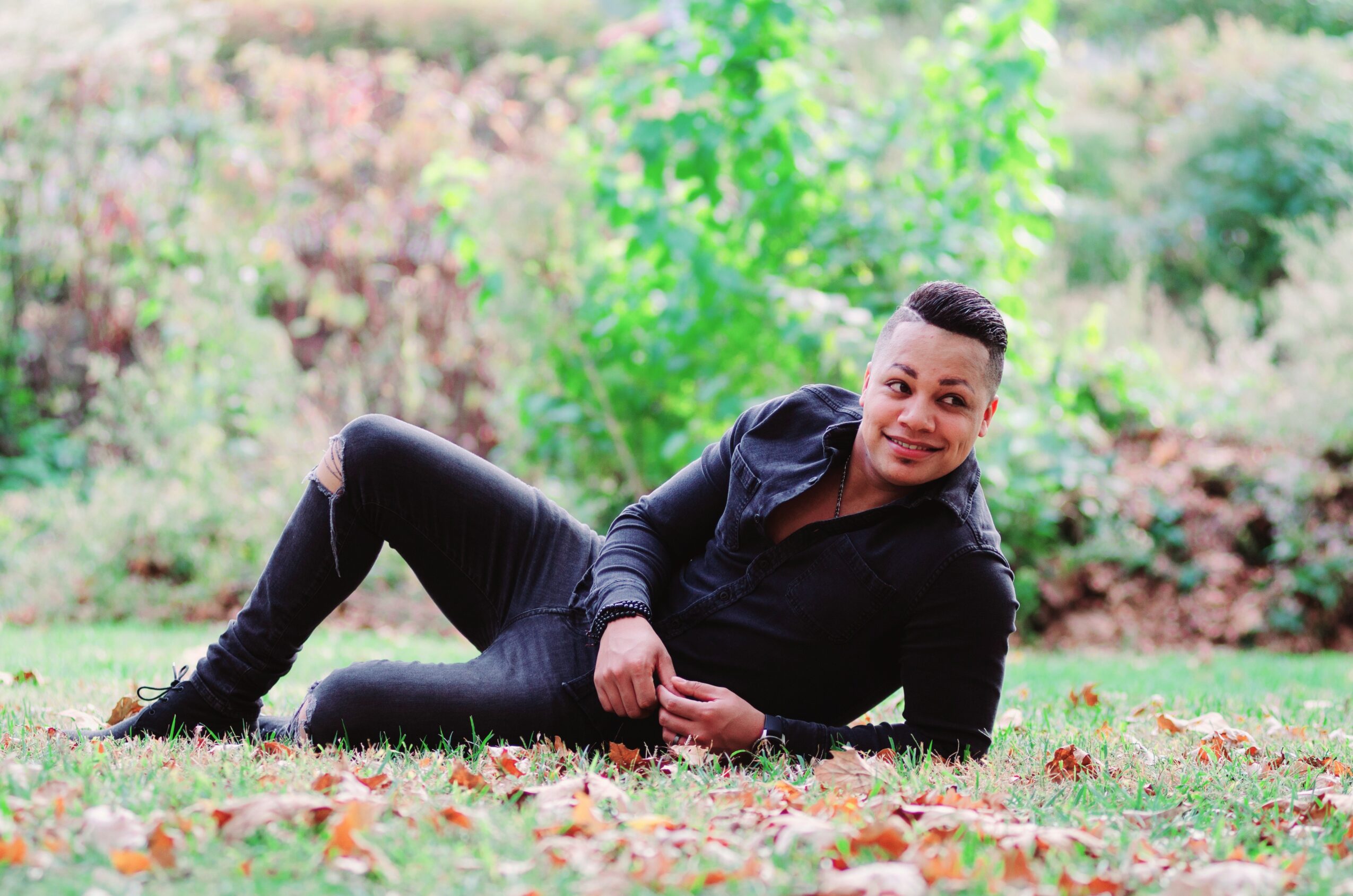 Person lying on grass in a park with trees and greenery in the background, smiling at the camera.