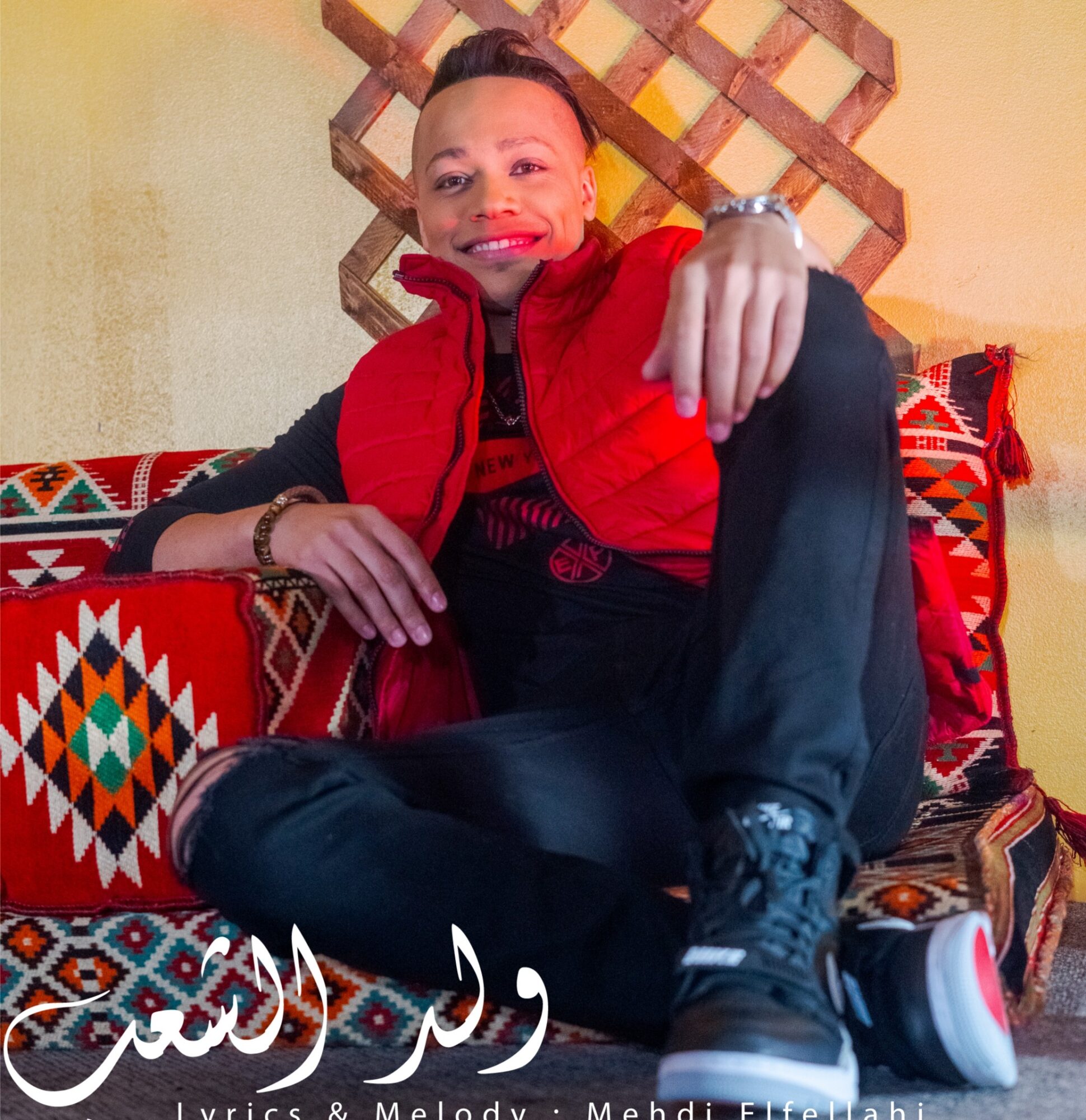 Person sitting on colorful patterned cushions, smiling, wearing a red vest and black pants, with a decorative wall behind.