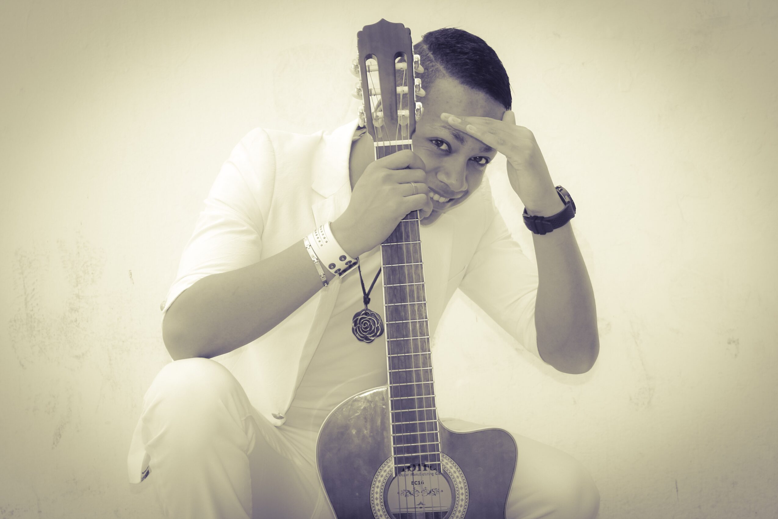 Person in white clothing holding a guitar, with one hand on forehead, against a plain background.