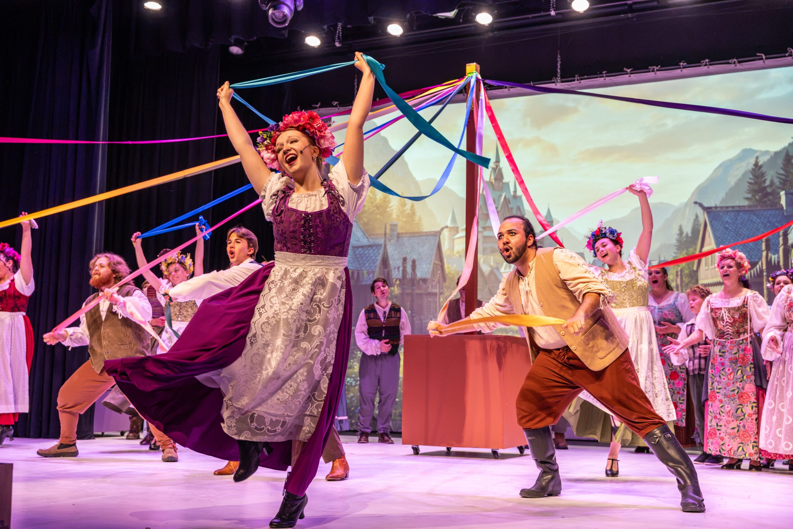 A lively stage scene with a woman dancing, surrounded by performers in colorful costumes, ribbons, and a scenic backdrop.