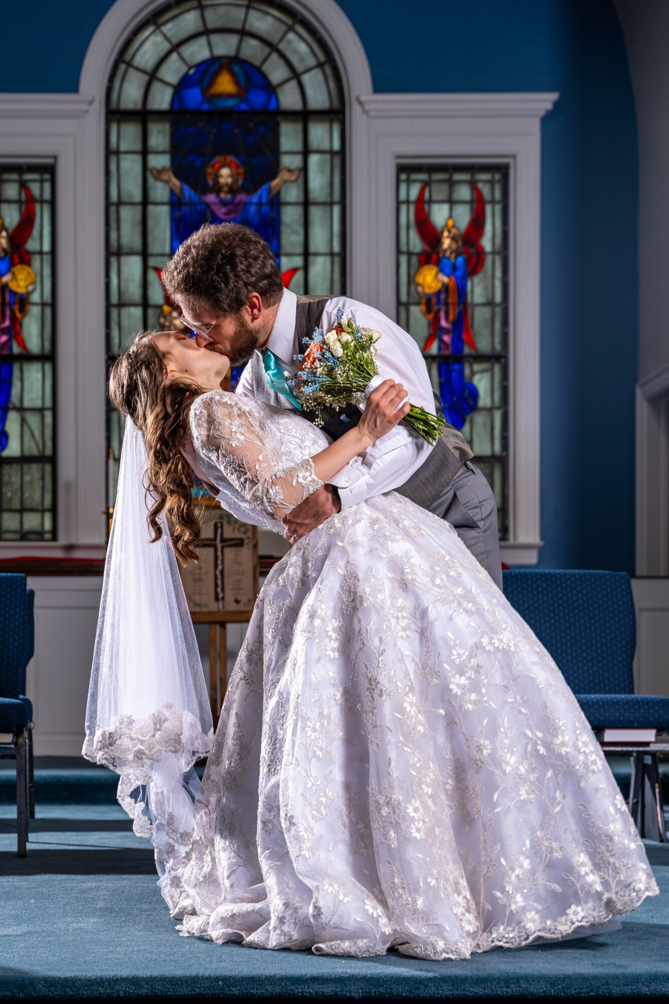 Couple dancing in a church with stained glass windows behind them, the woman in a white wedding dress and the man in a suit