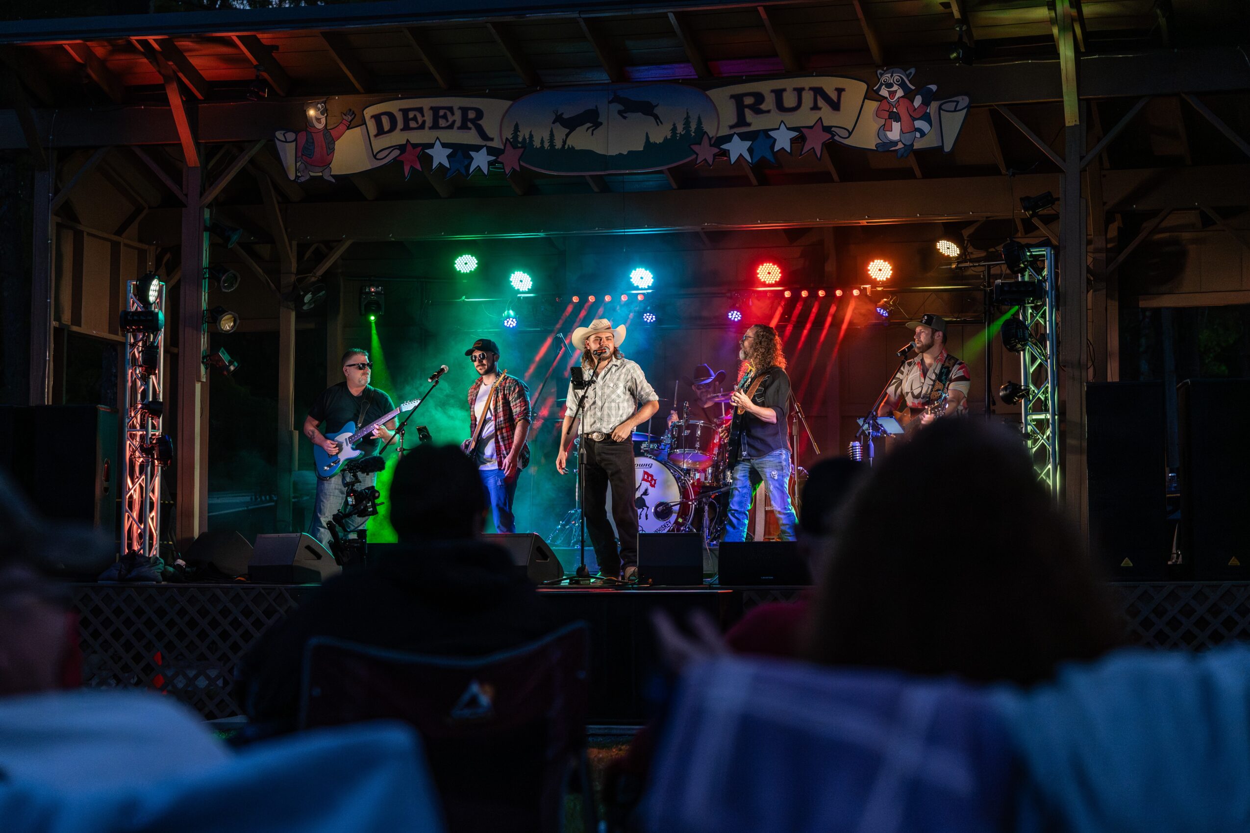 Band performs on stage with colorful lights, audience in foreground, and a banner reading 'Deer Run' above.