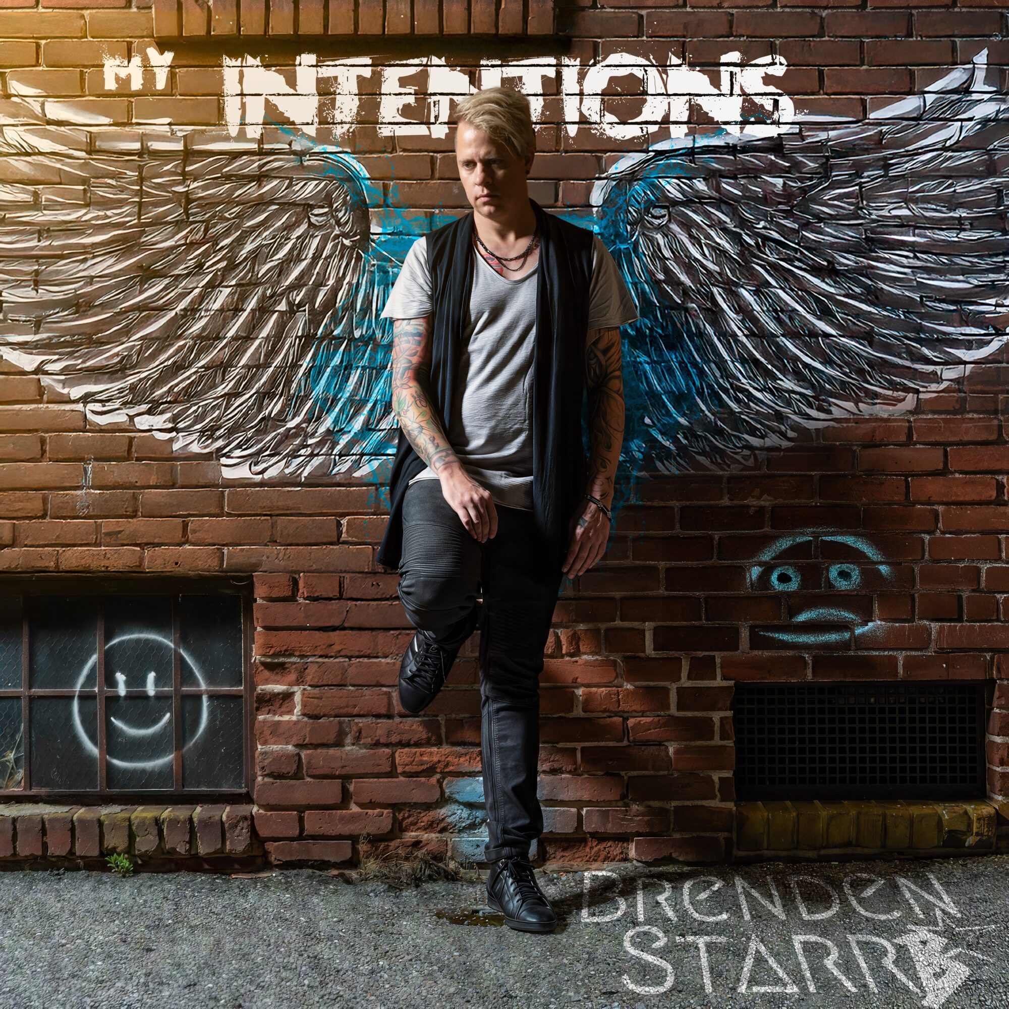 Young man with wings painted on brick wall behind him, standing outdoors, wearing a backpack and casual clothing.