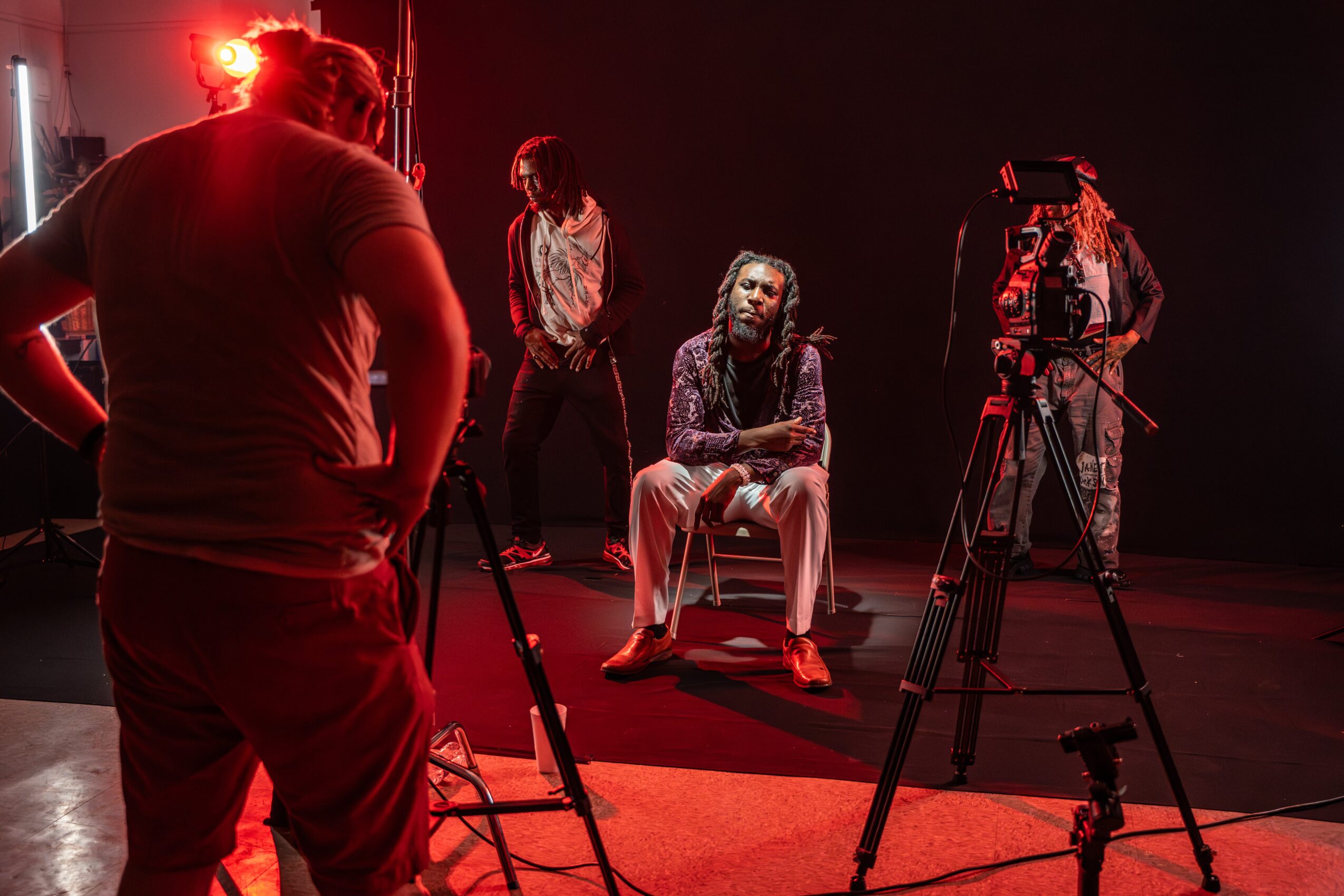 Person sitting on a chair in a dark studio with three people and camera equipment, illuminated by red lighting.