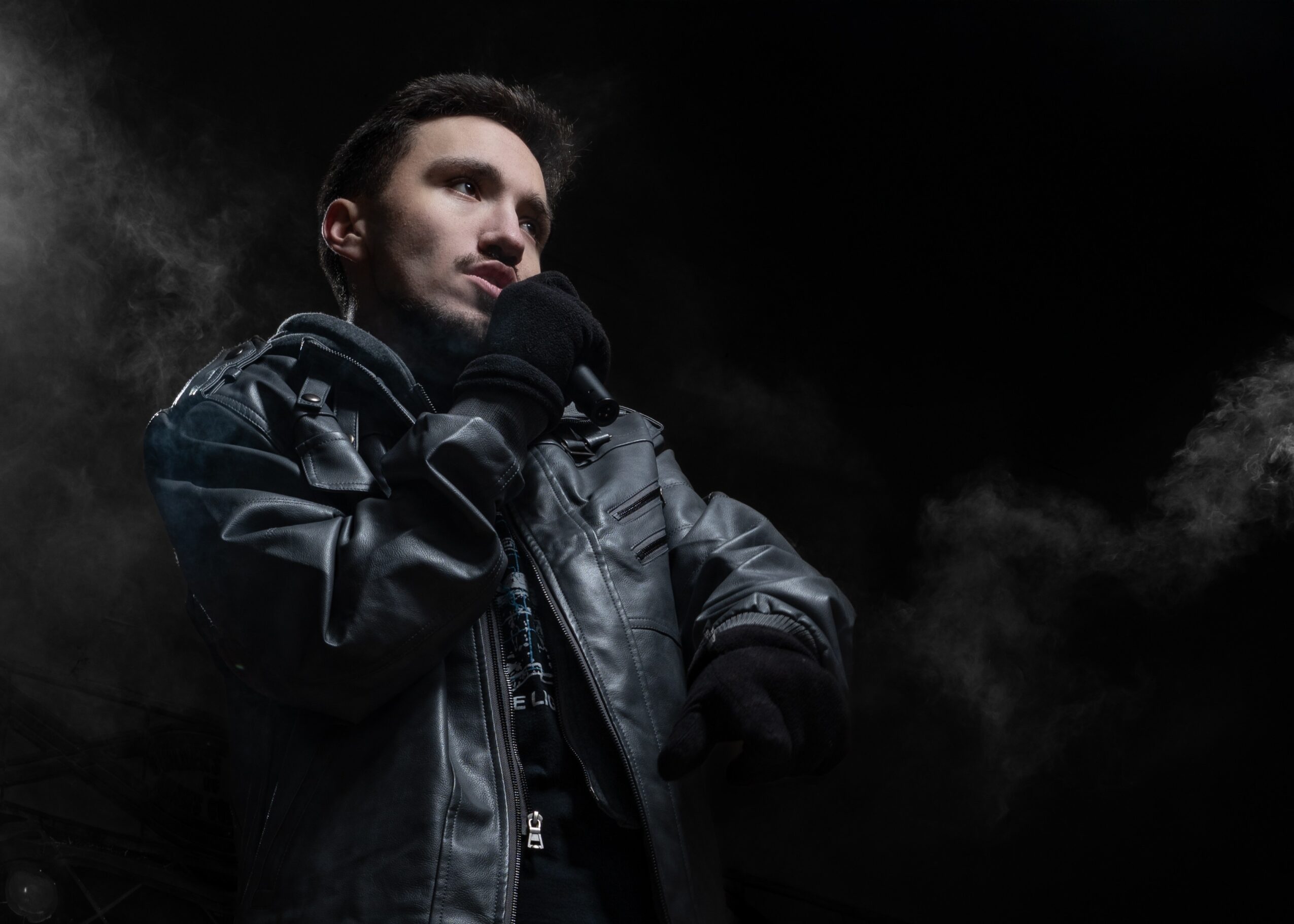 Young man in black leather jacket and gloves, looking thoughtfully into the distance against a dark background.