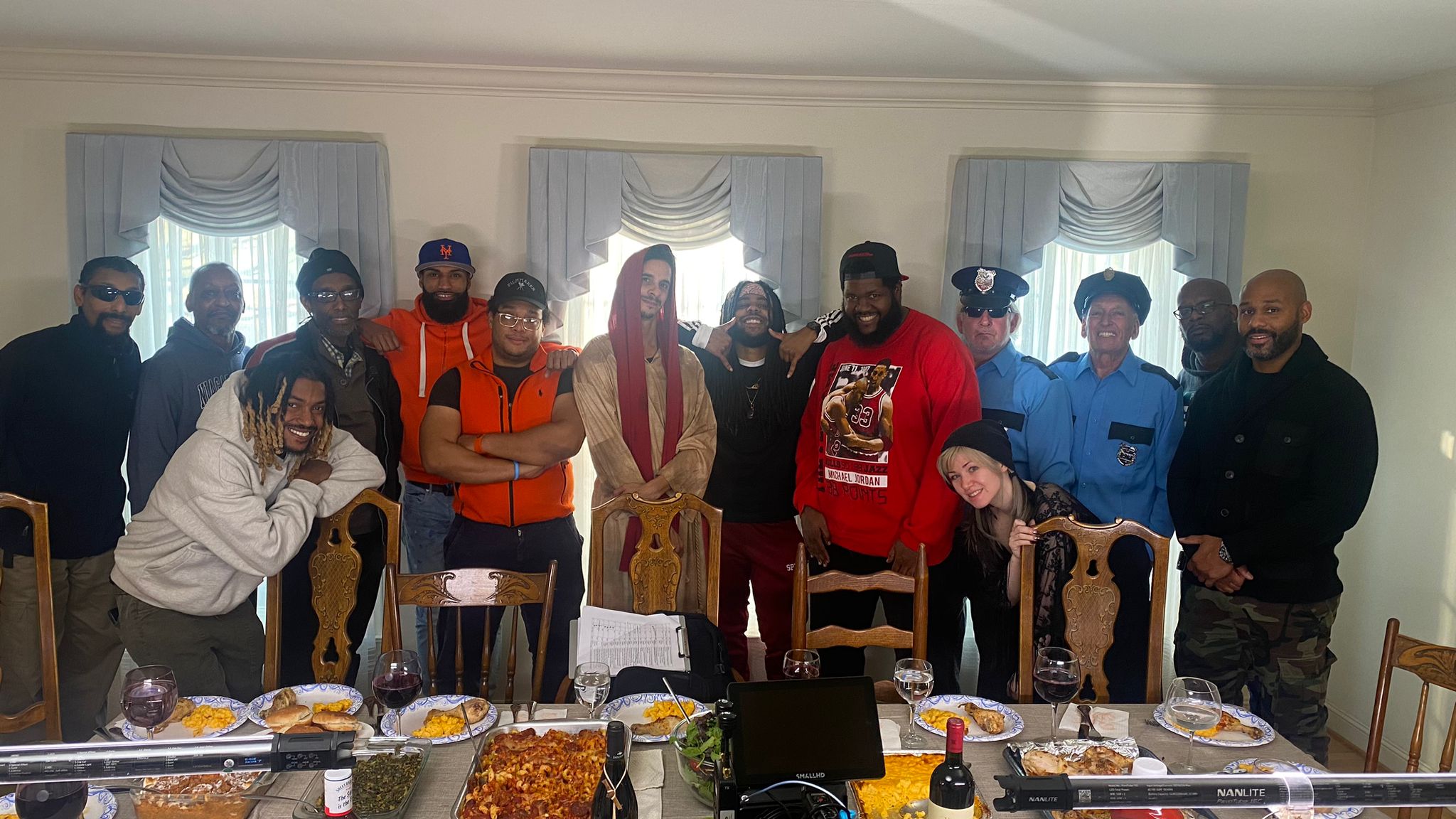 Group of people standing behind a table with food and drinks in a room with curtains and windows.