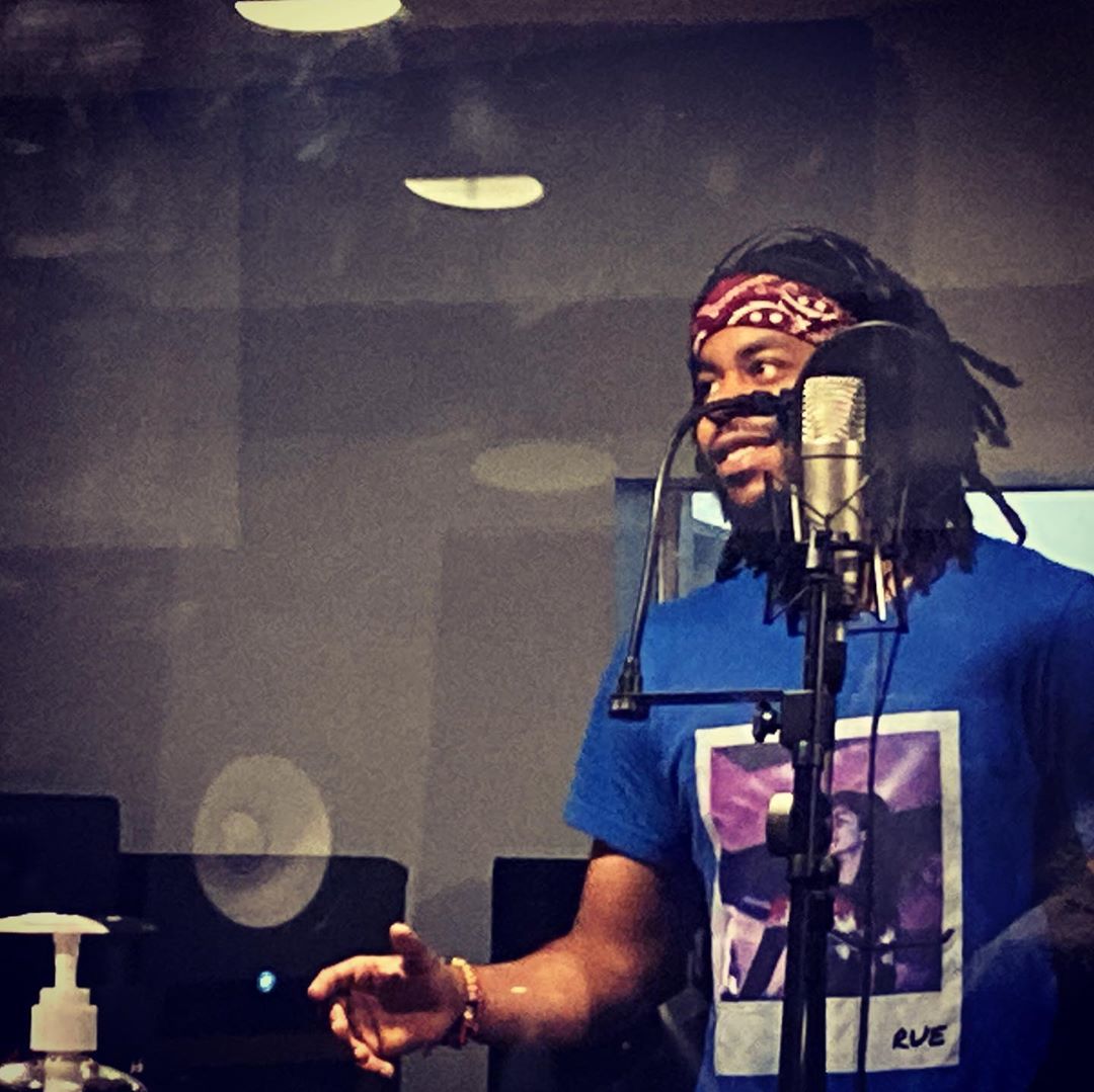 Person with dreadlocks and headband recording in a studio, wearing a blue shirt, microphone in front, glass window reflection.