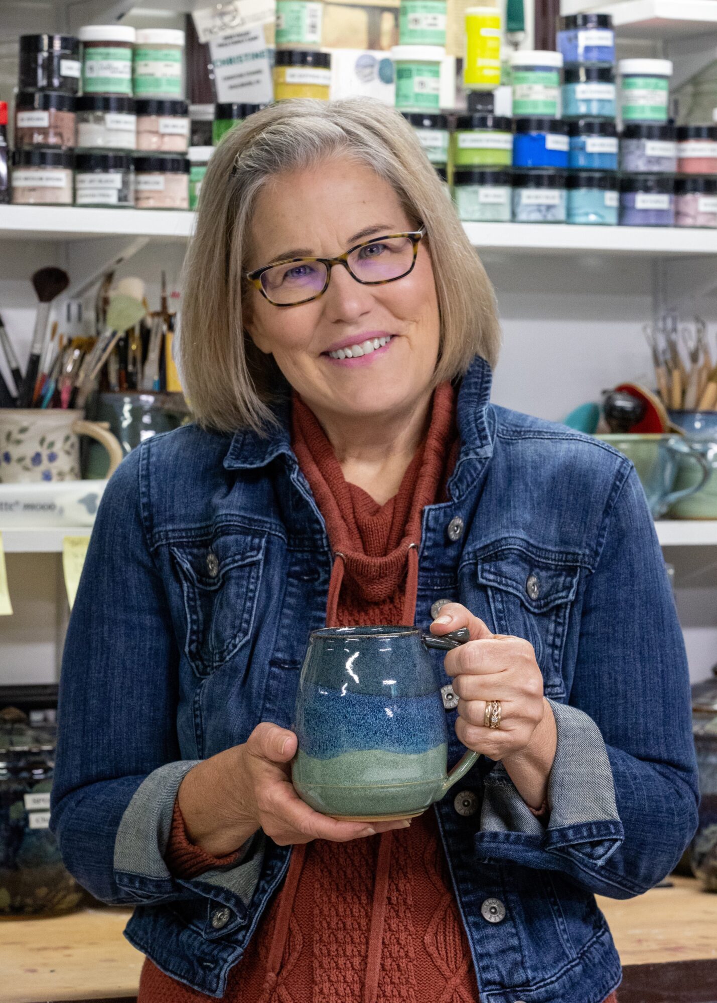 Woman with glasses and shoulder-length hair holding a ceramic mug, standing in an art supplies store.