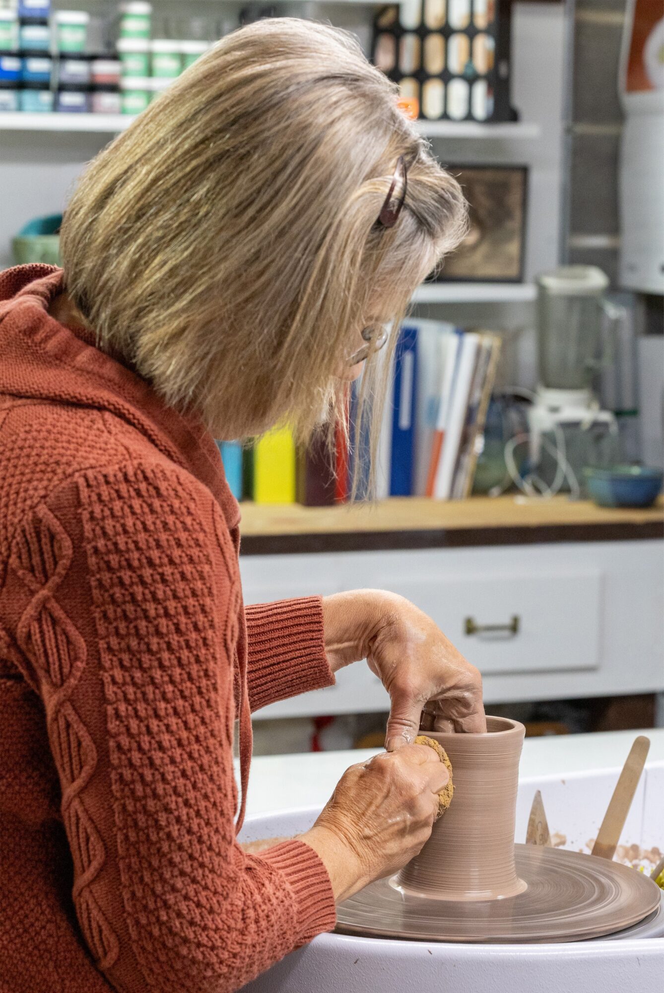 Woman with gray hair working on a pottery wheel in a studio, wearing a red sweater, with shelves and tools in background.