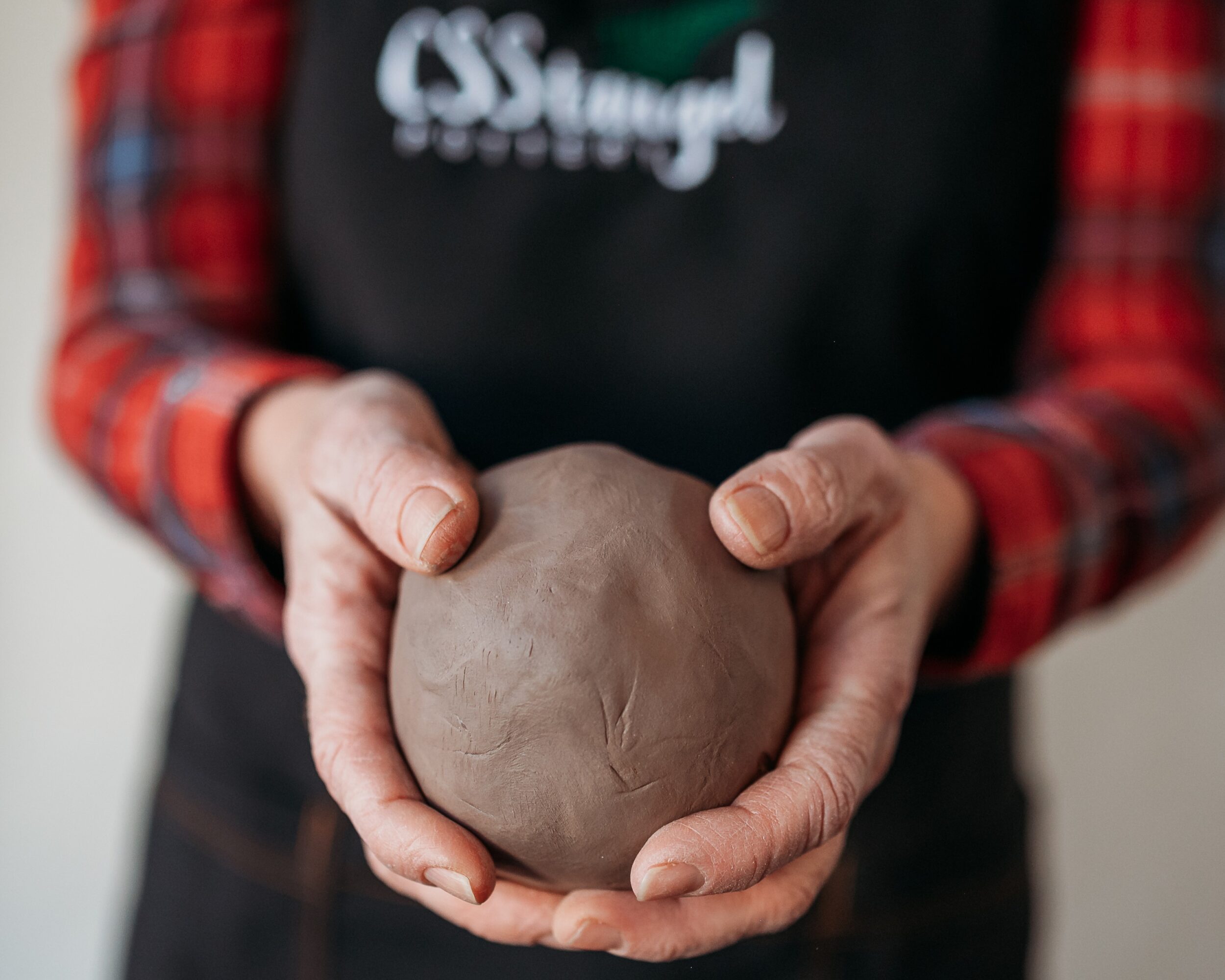 Person holding a ball of clay with both hands, wearing a black apron and red plaid shirt.
