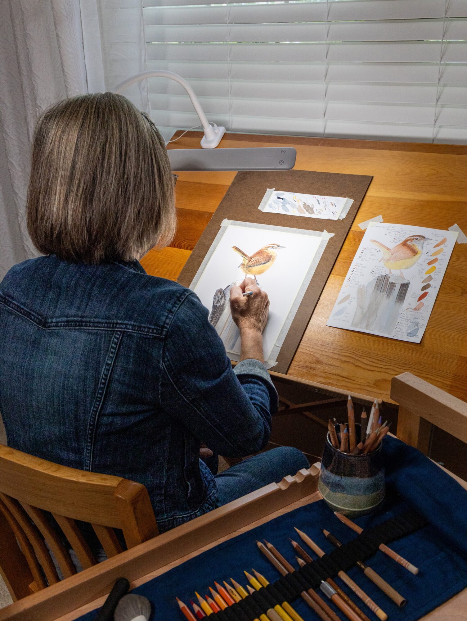 Person drawing on a digital tablet at a wooden desk with papers and art supplies, window with blinds in background.