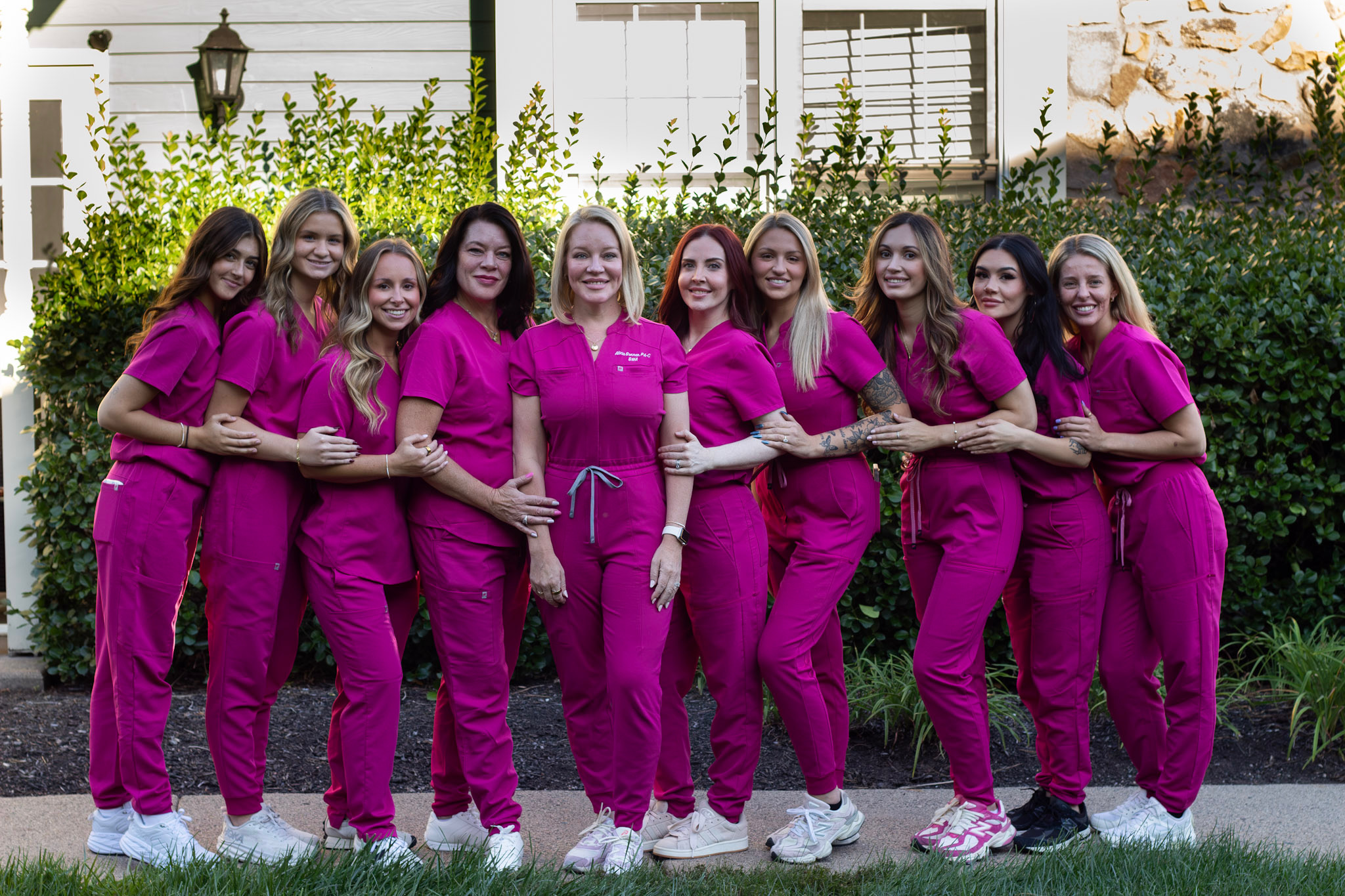 Group of nine women in matching pink scrubs standing outdoors in front of a house and greenery, smiling, holding hands.