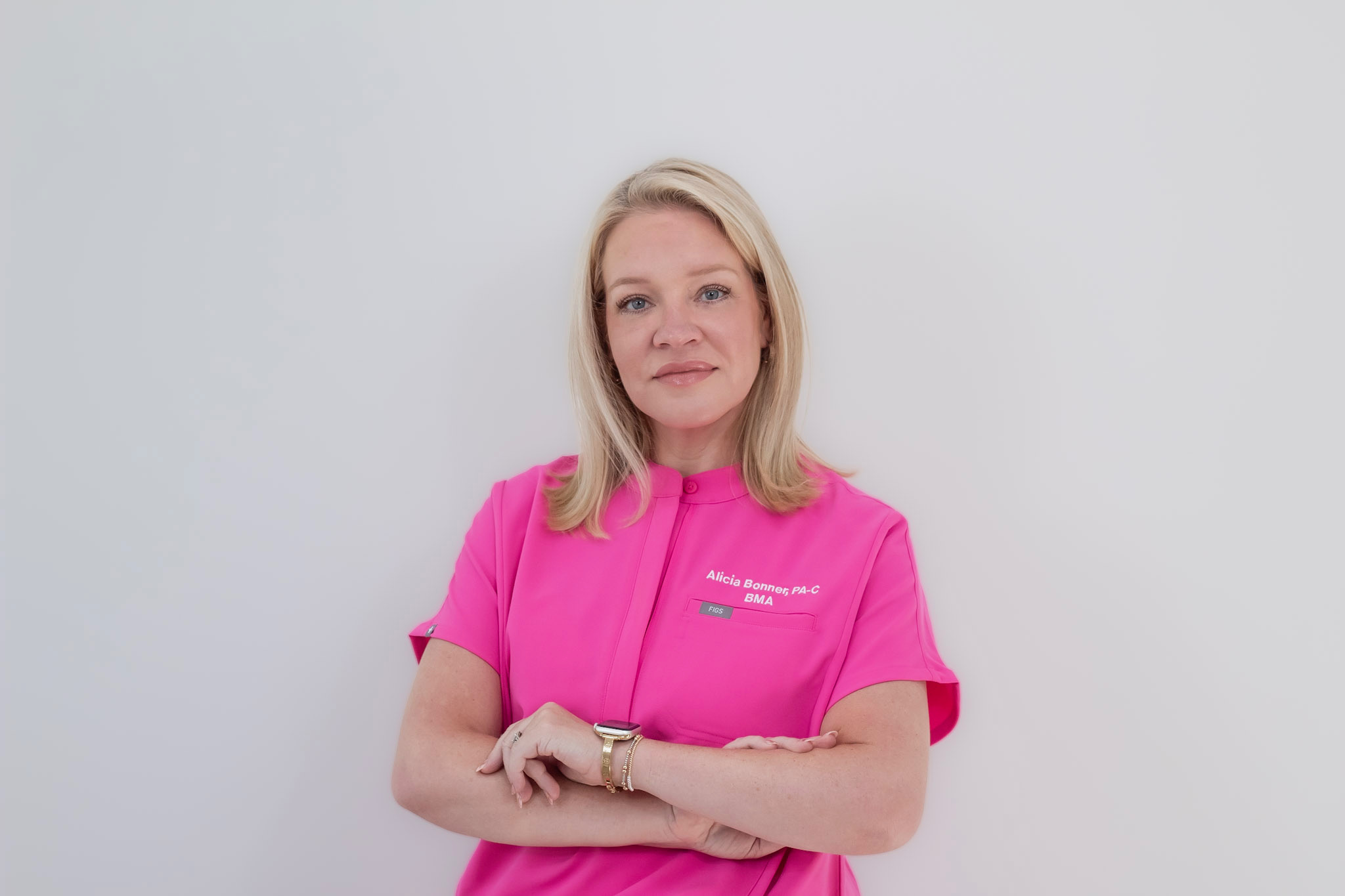 Woman with blonde hair wearing a pink shirt, standing against a plain light background, arms crossed.
