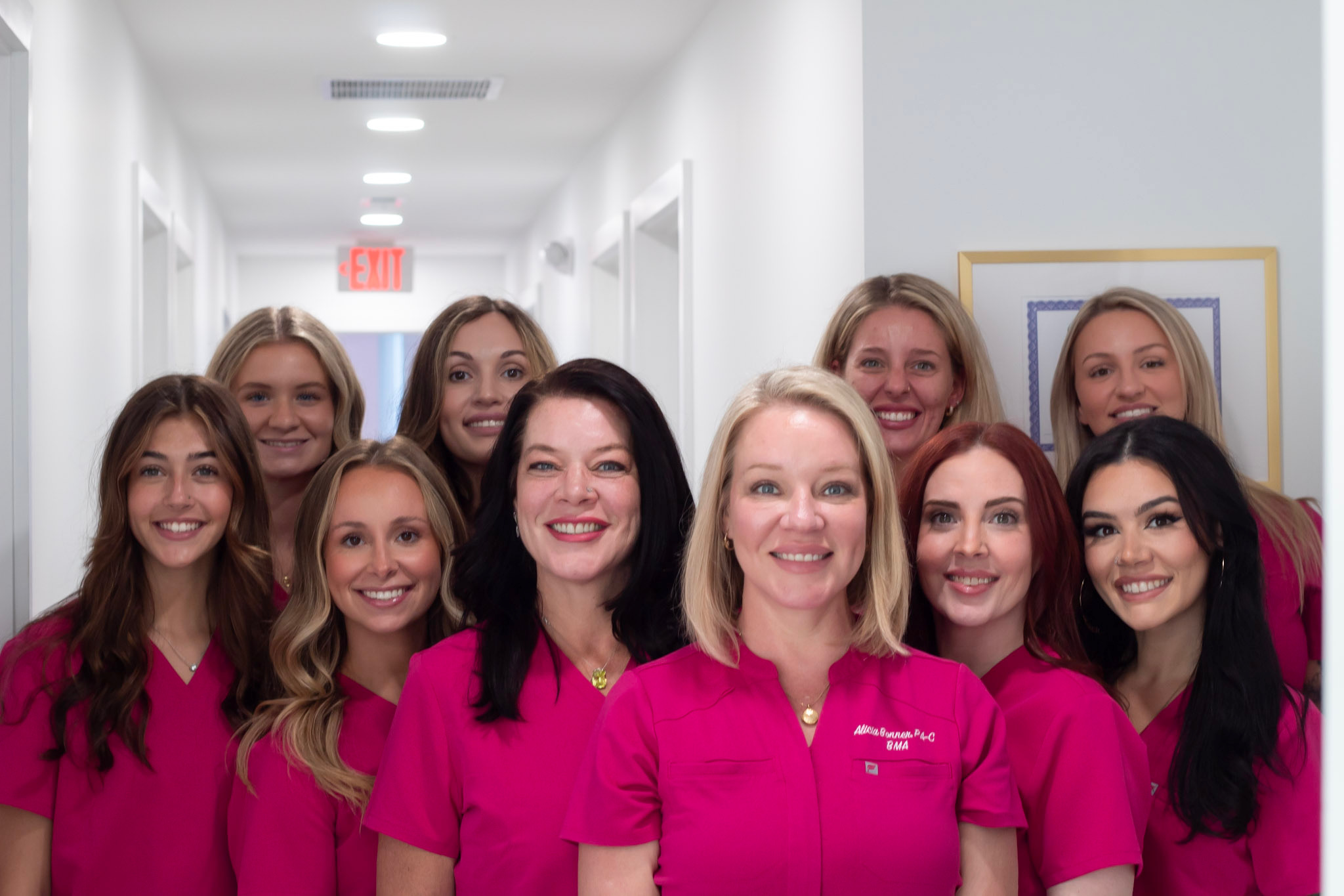 Group of ten women in pink uniforms smiling in a hallway with white walls and doors.