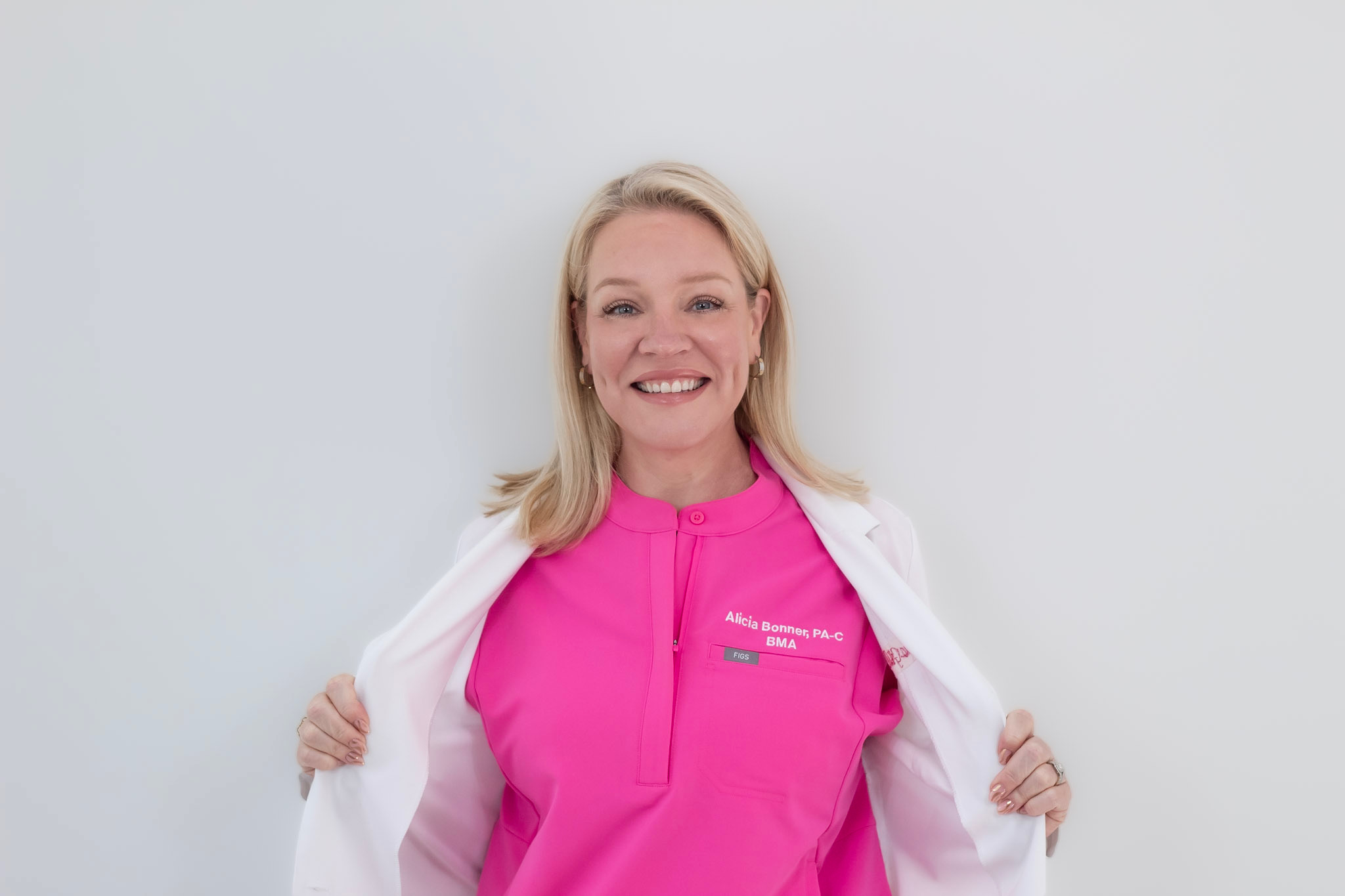 Smiling woman with blonde hair wearing a pink top and white jacket, standing against a plain background.