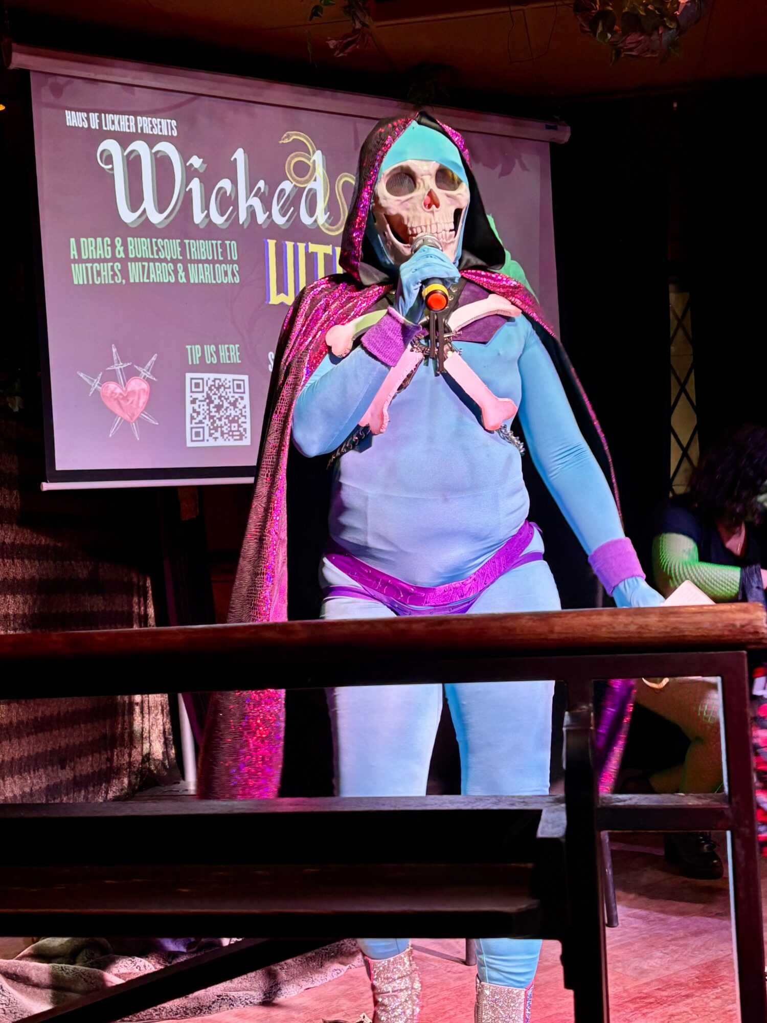 Person in a skeleton costume with a skull mask, wearing a cape, speaking into a microphone on stage.