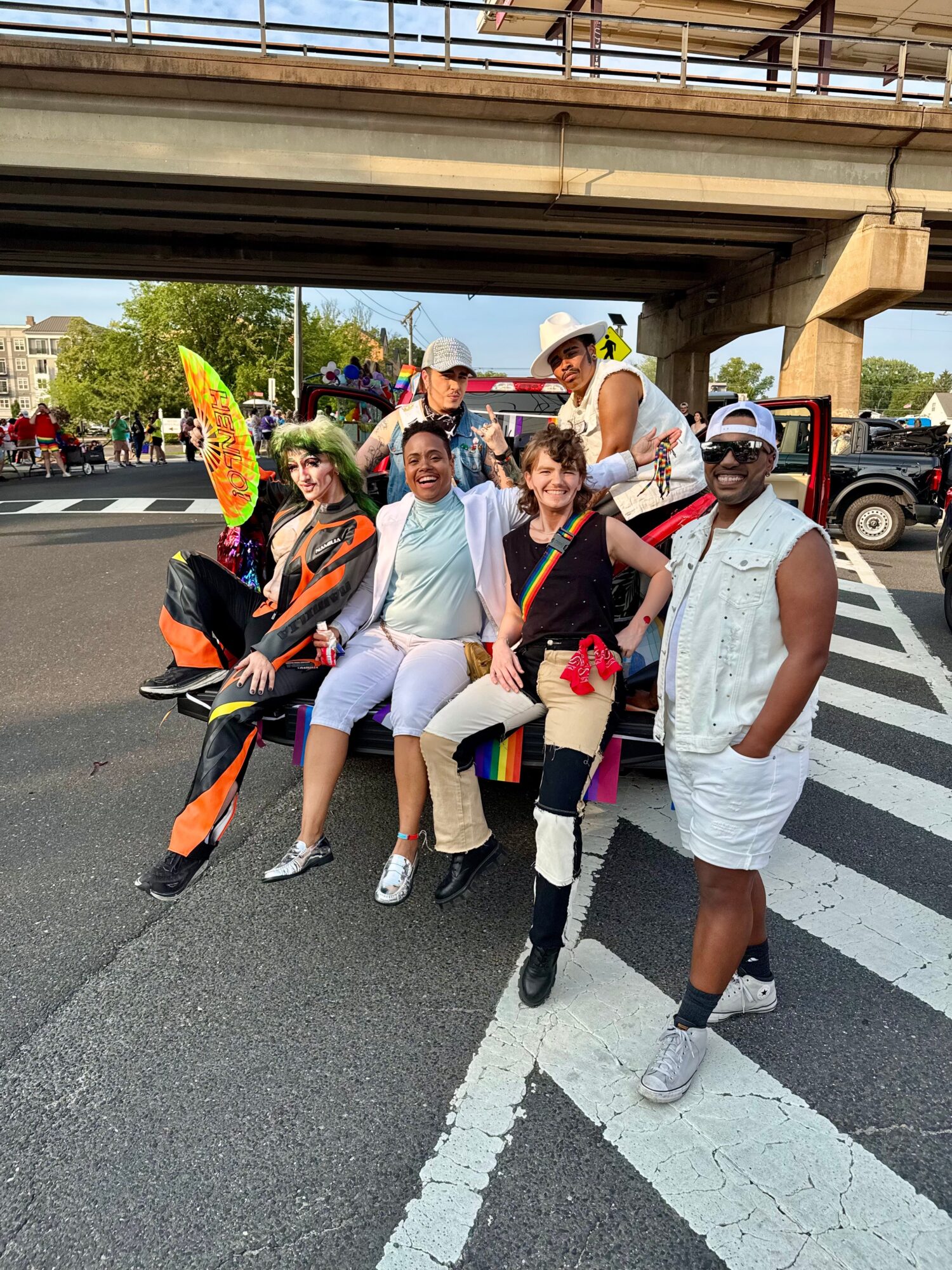 Group of six people posing under a bridge, some in colorful costumes, on a street with cars and trees in background.