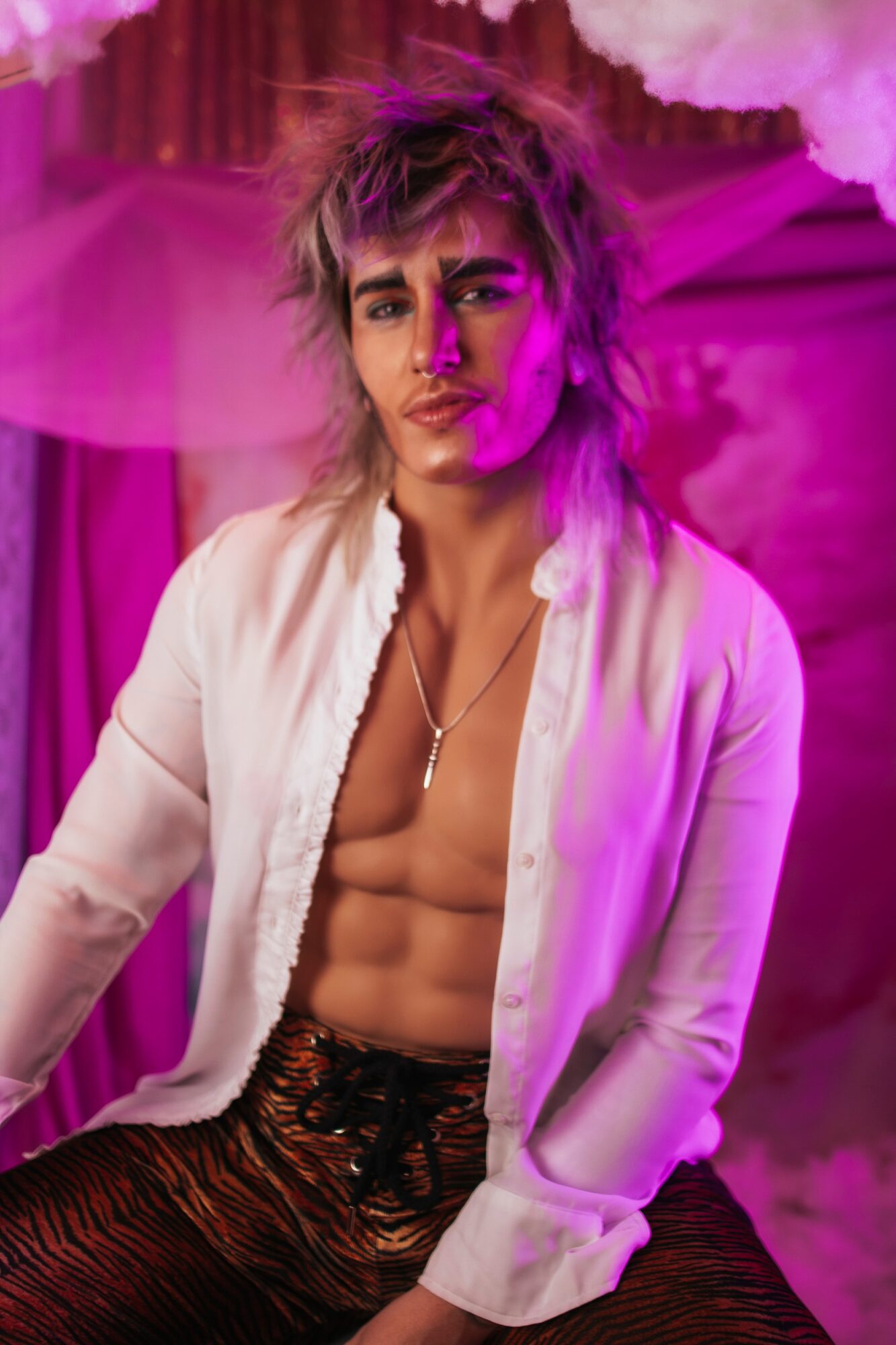 Young man with styled hair, shirt open showing abs, sitting in a pink-lit room with curtains and clouds, looking at camera.