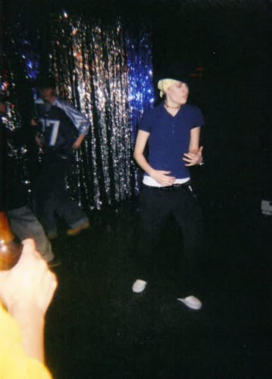 Person with blonde hair in a blue shirt and black pants standing on stage with a glittery backdrop.