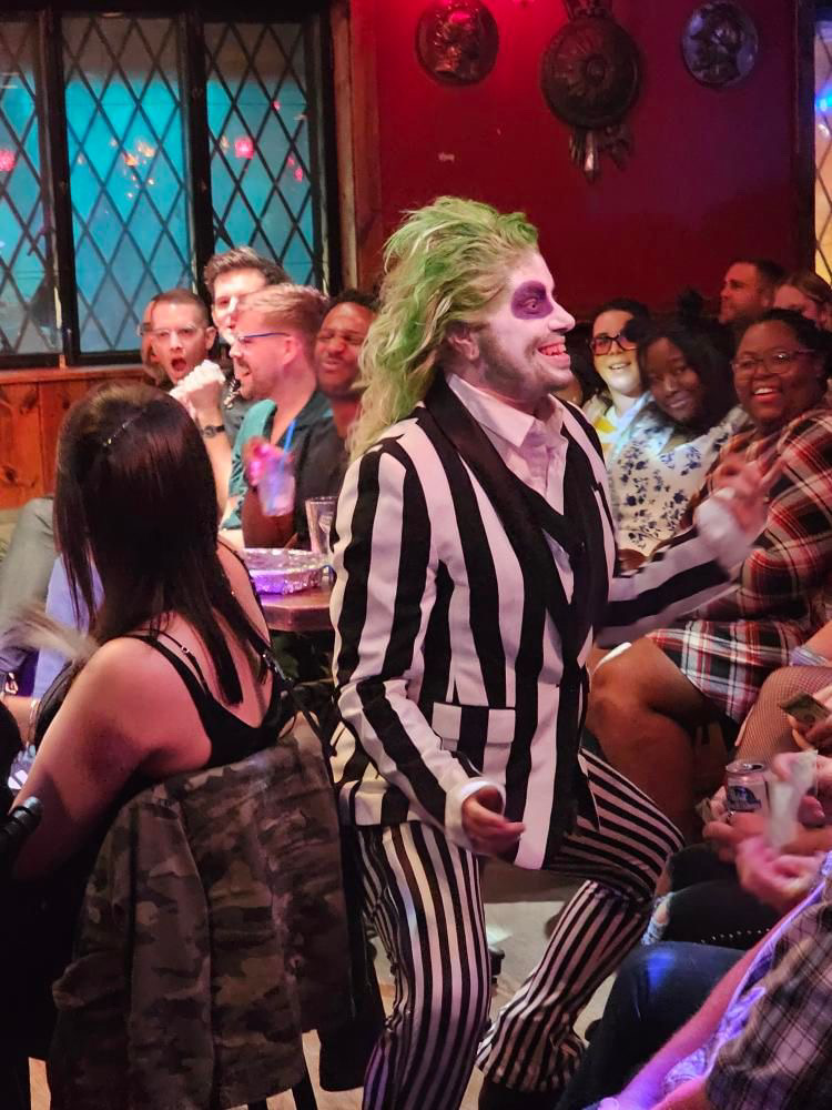 Person dressed as the Joker with green hair, striped suit, and face paint, standing among seated audience members in a decorated room.