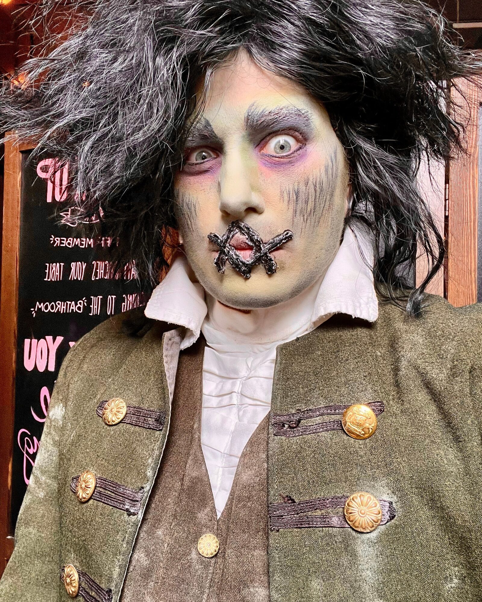 Person with theatrical makeup, wild hair, and vintage military-style jacket, looking directly at the camera.