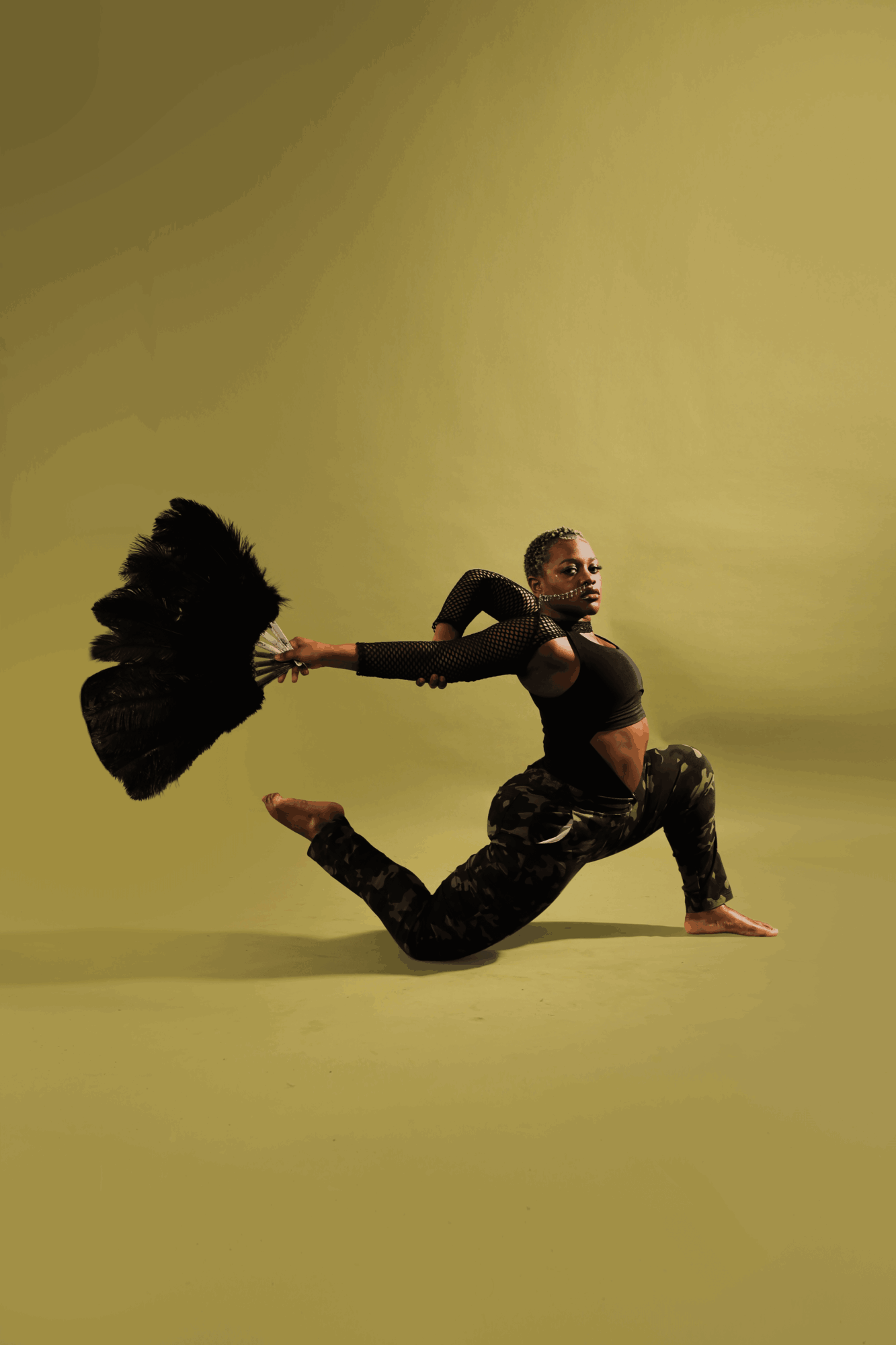 Person in black top and camouflage pants kneels on one knee, holding a large black feathered fan, against a yellow background.