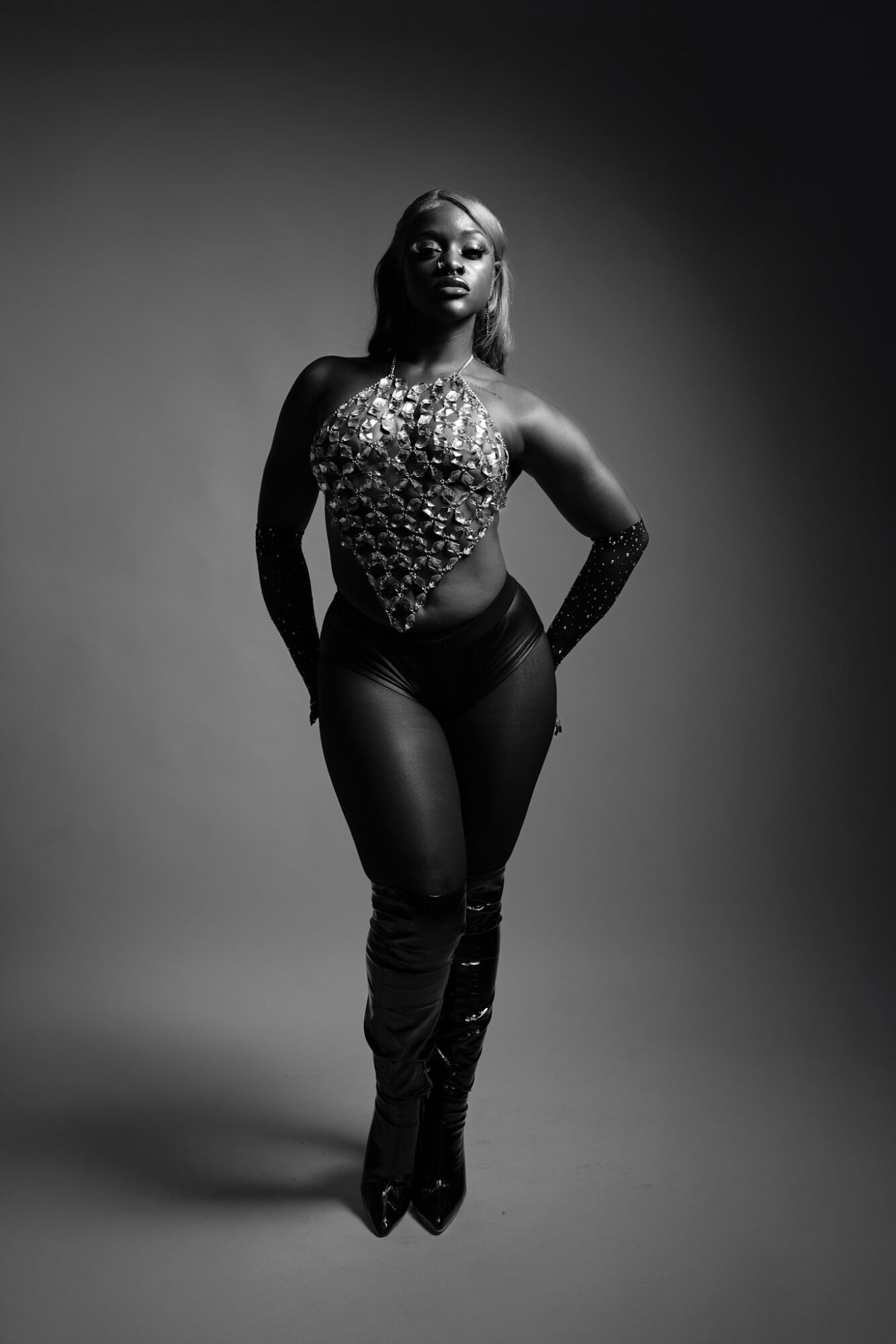 Woman in a fitted outfit with a patterned top and high-waisted pants, standing with hands on hips, in black and white.