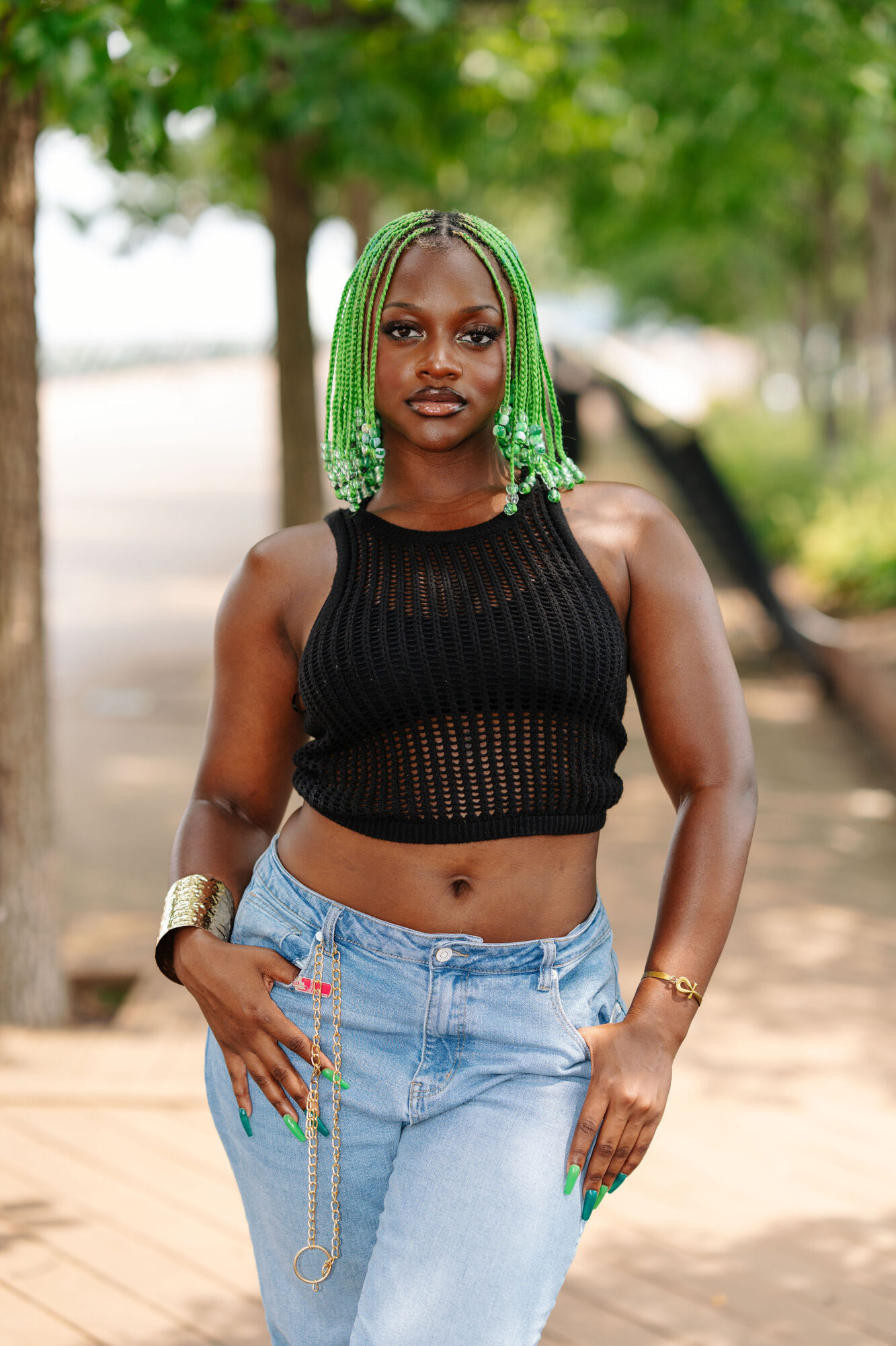 Woman with green braided hair wearing a black sleeveless top and light blue jeans outdoors, trees in background.