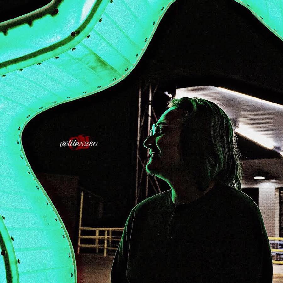 Person with shoulder-length hair smiling in silhouette against a brightly lit, curved, green structure.