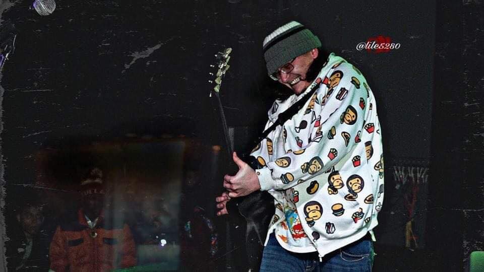 Person wearing a patterned jacket and beanie playing guitar outdoors at night.