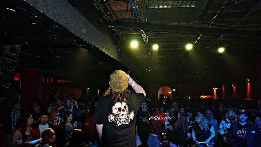 Person with a hat and skull shirt singing into a microphone on stage, audience watching in a dark venue.