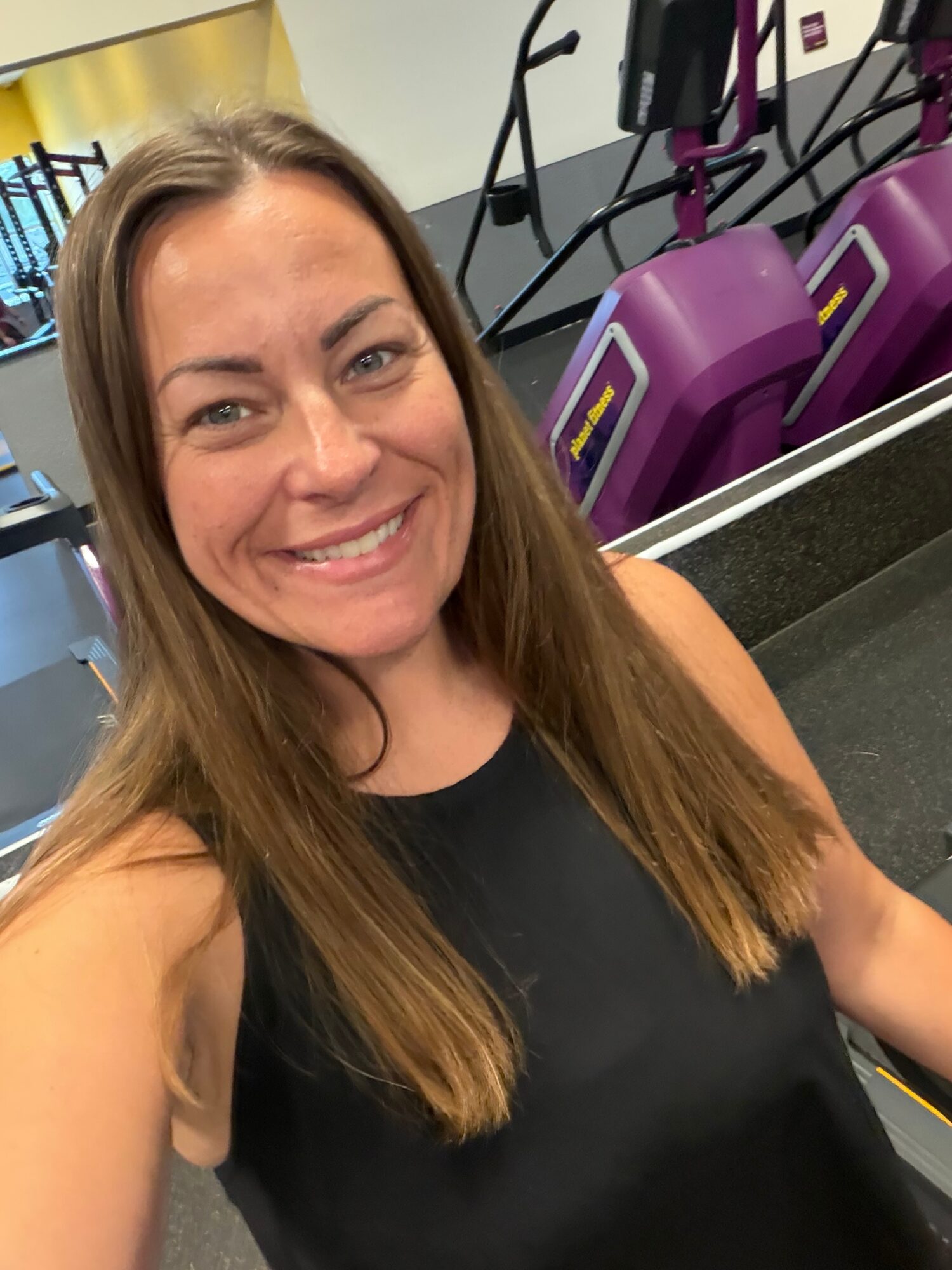 Smiling woman with long hair in a gym, with exercise equipment in the background.