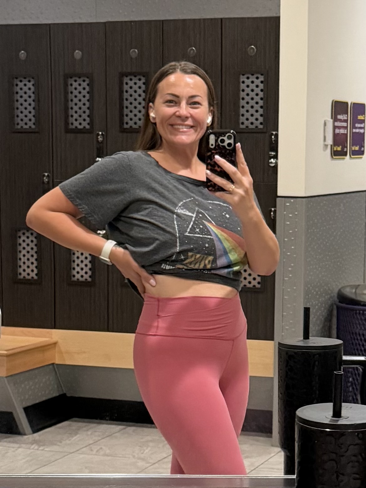 Smiling woman taking a mirror selfie in a gym locker room, wearing a gray crop top and pink leggings.