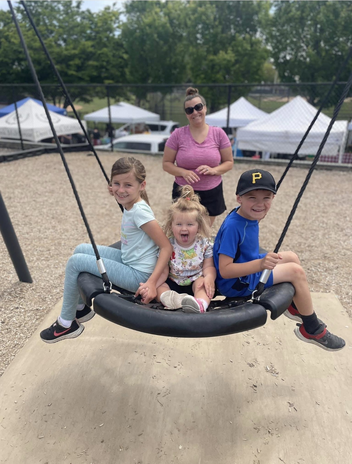 Three children sitting on a swing with a woman standing behind them outdoors, tents and trees in background.