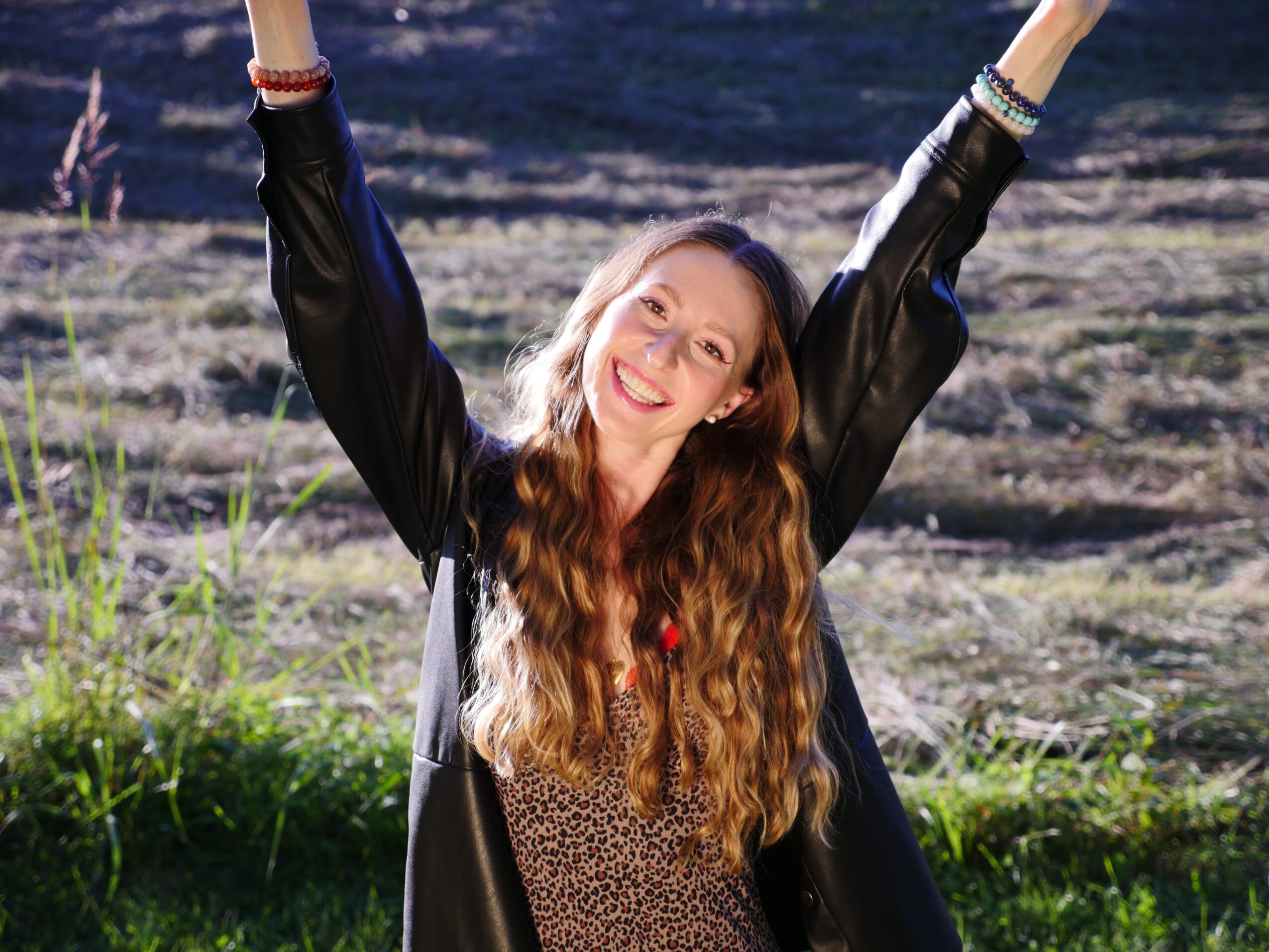 Young woman with long wavy hair smiling outdoors with arms raised, wearing a black jacket and patterned top.