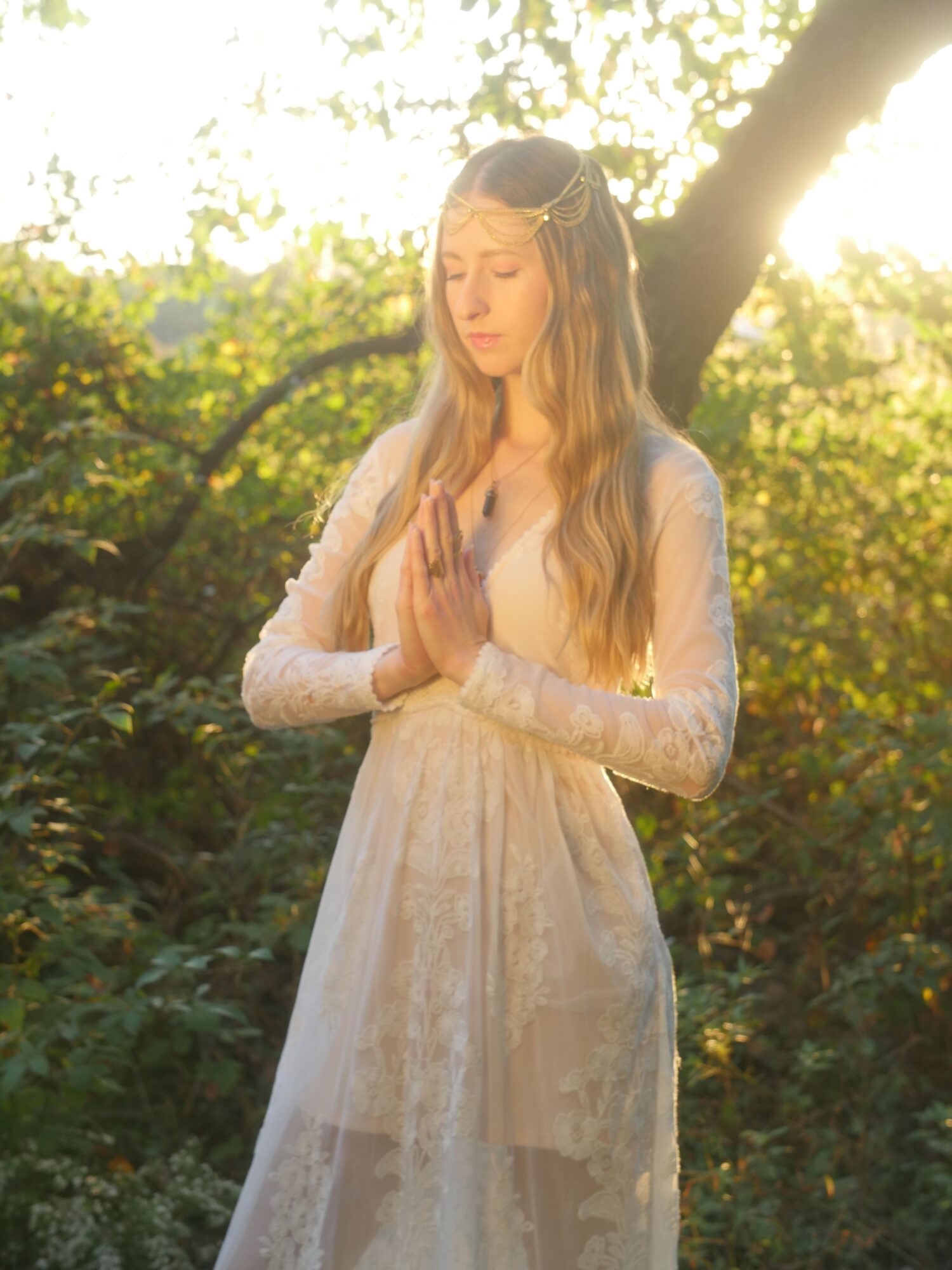 Woman in a white dress standing outdoors with trees and sunlight, holding her hands near her face.