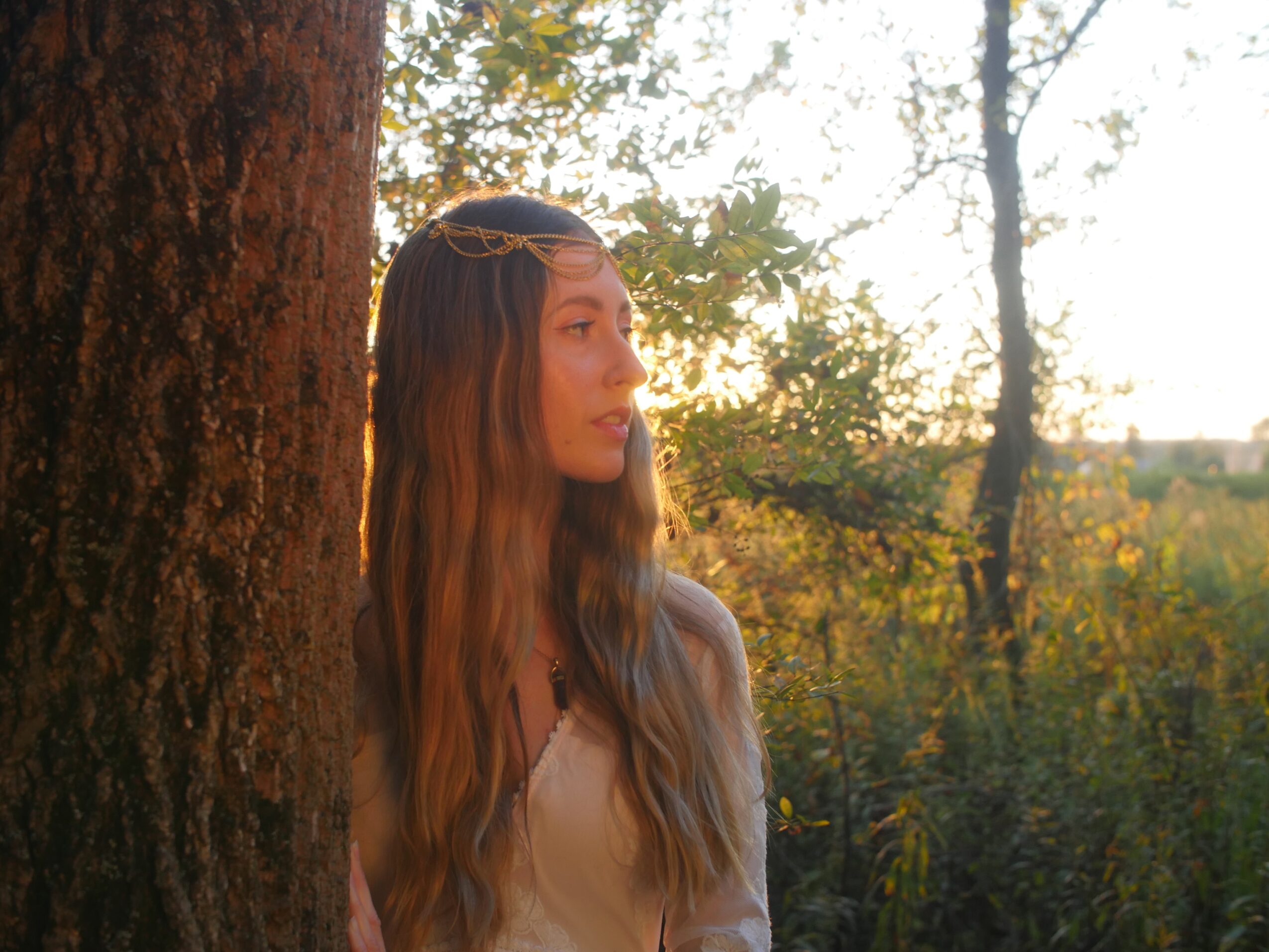 Young woman with long wavy hair standing outdoors near a tree during sunset, looking to the side.