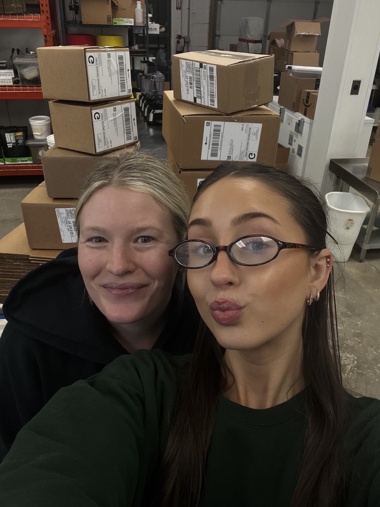 Two women taking a selfie in a warehouse with stacked boxes and shelves in the background.