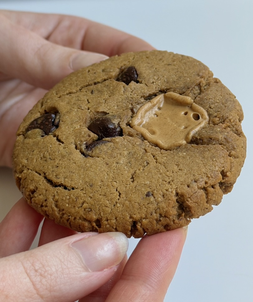Person holding a large chocolate chip cookie with a peanut butter cup in the center, against a light background.