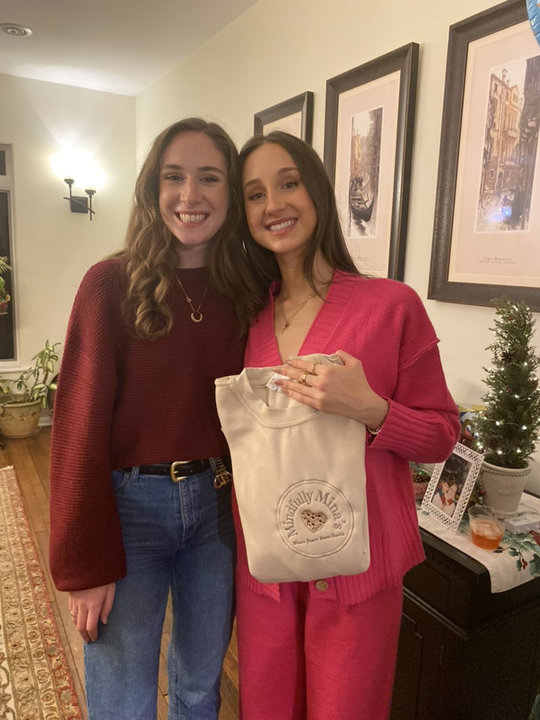 Two women smiling indoors, one holding a beige tote bag, standing in front of framed pictures and a small Christmas tree.