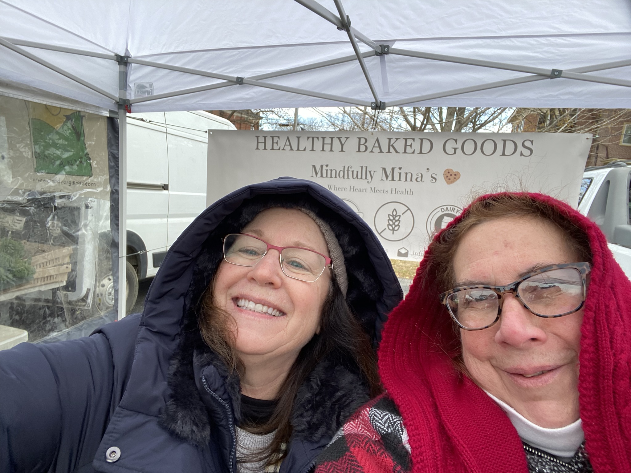 Two women smiling under a canopy, one wearing glasses and a dark hood, the other with glasses and a red hood, in front of a sign that reads 'Healthy Baked Goods'.
