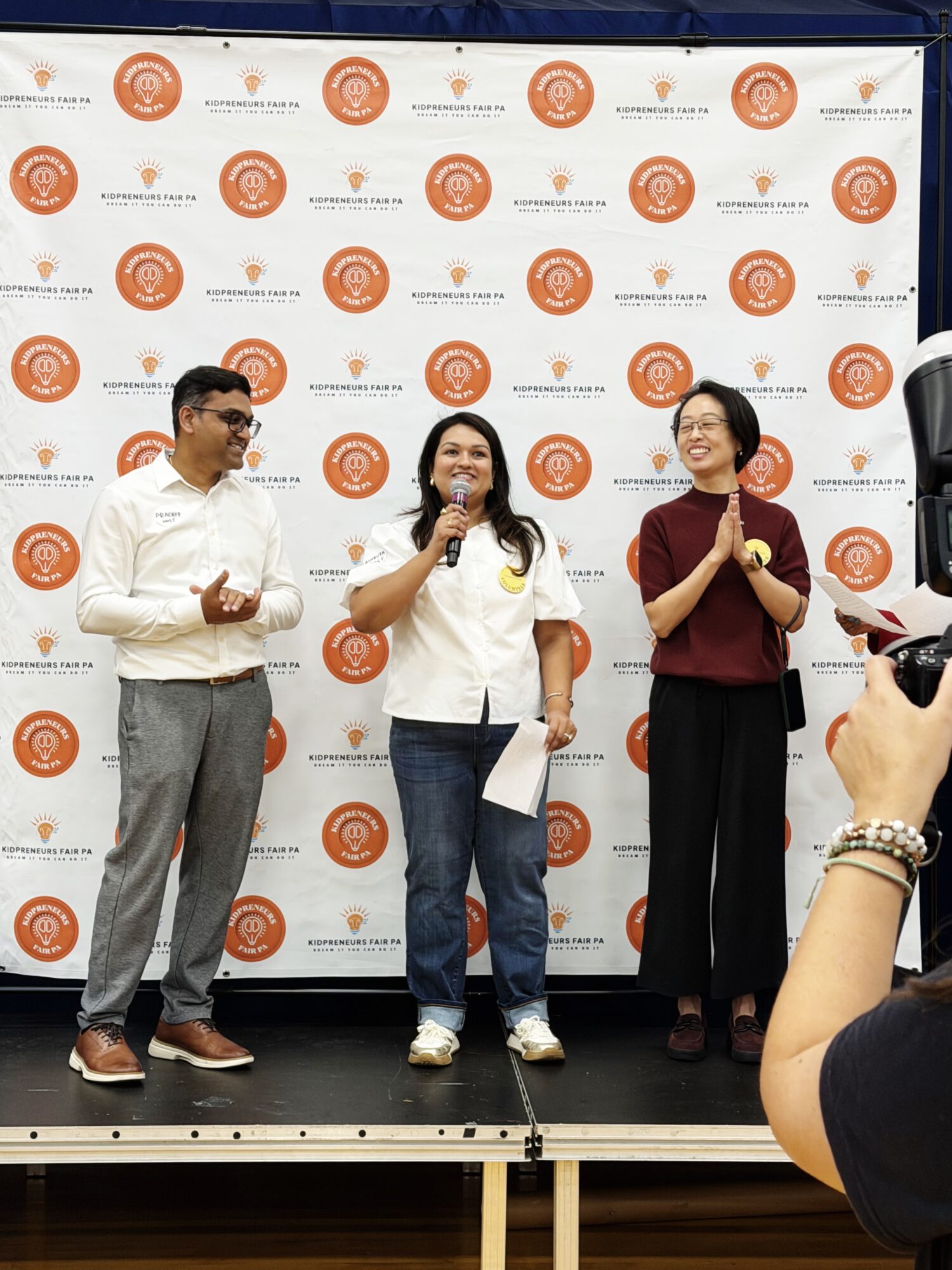 Three people standing on a stage in front of a backdrop with orange logos, one woman speaking into a microphone, others clapping.