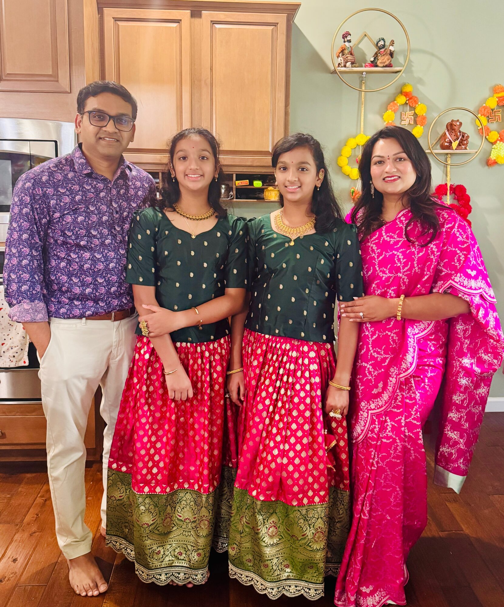 Family of four standing in a kitchen, two girls in traditional dresses, man in glasses, woman in pink saree, decorated for festival