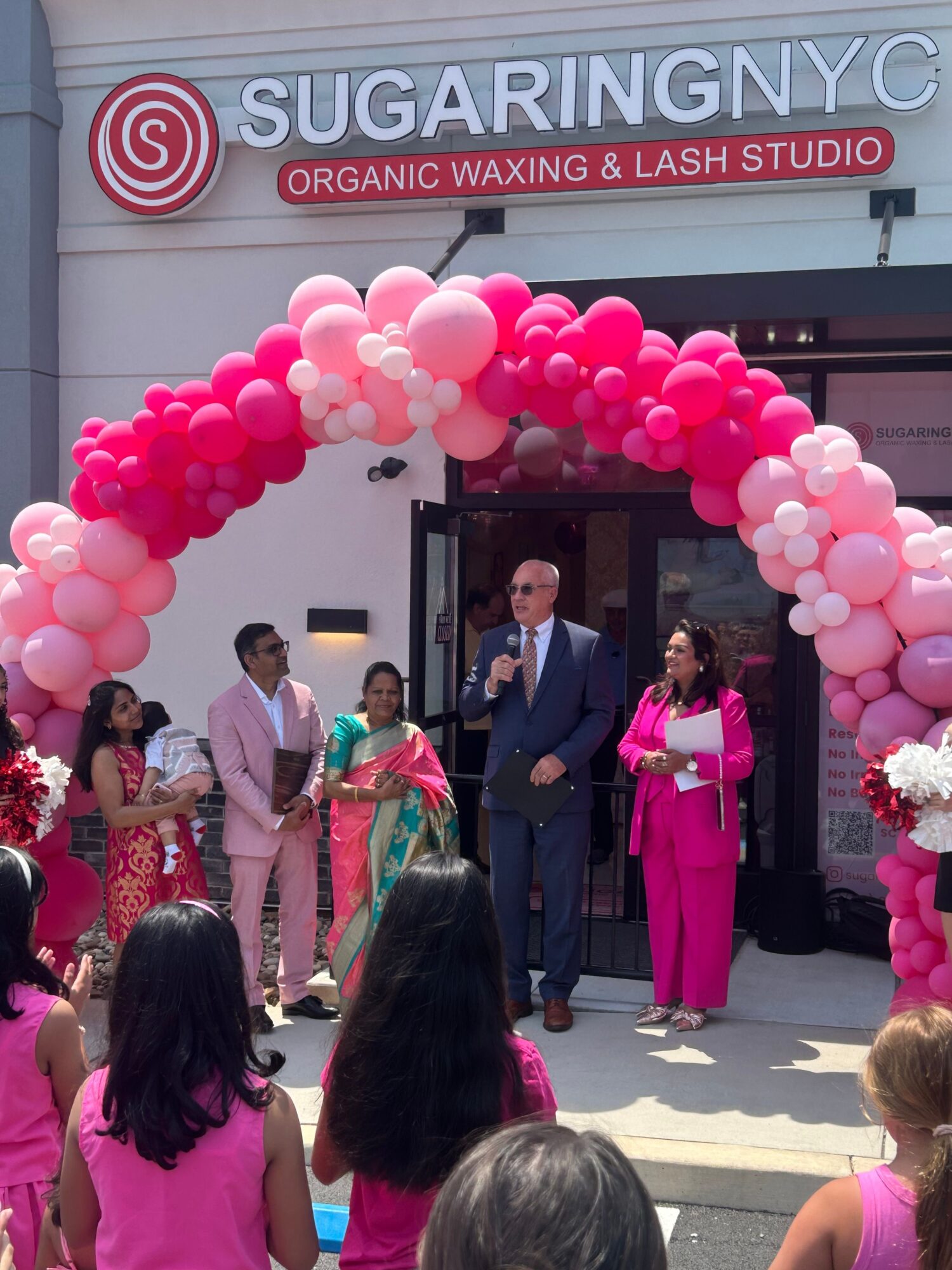 People gather outside a salon with a pink balloon arch, celebrating an event, with a sign reading 'Sugaring & Lashing Studio'.